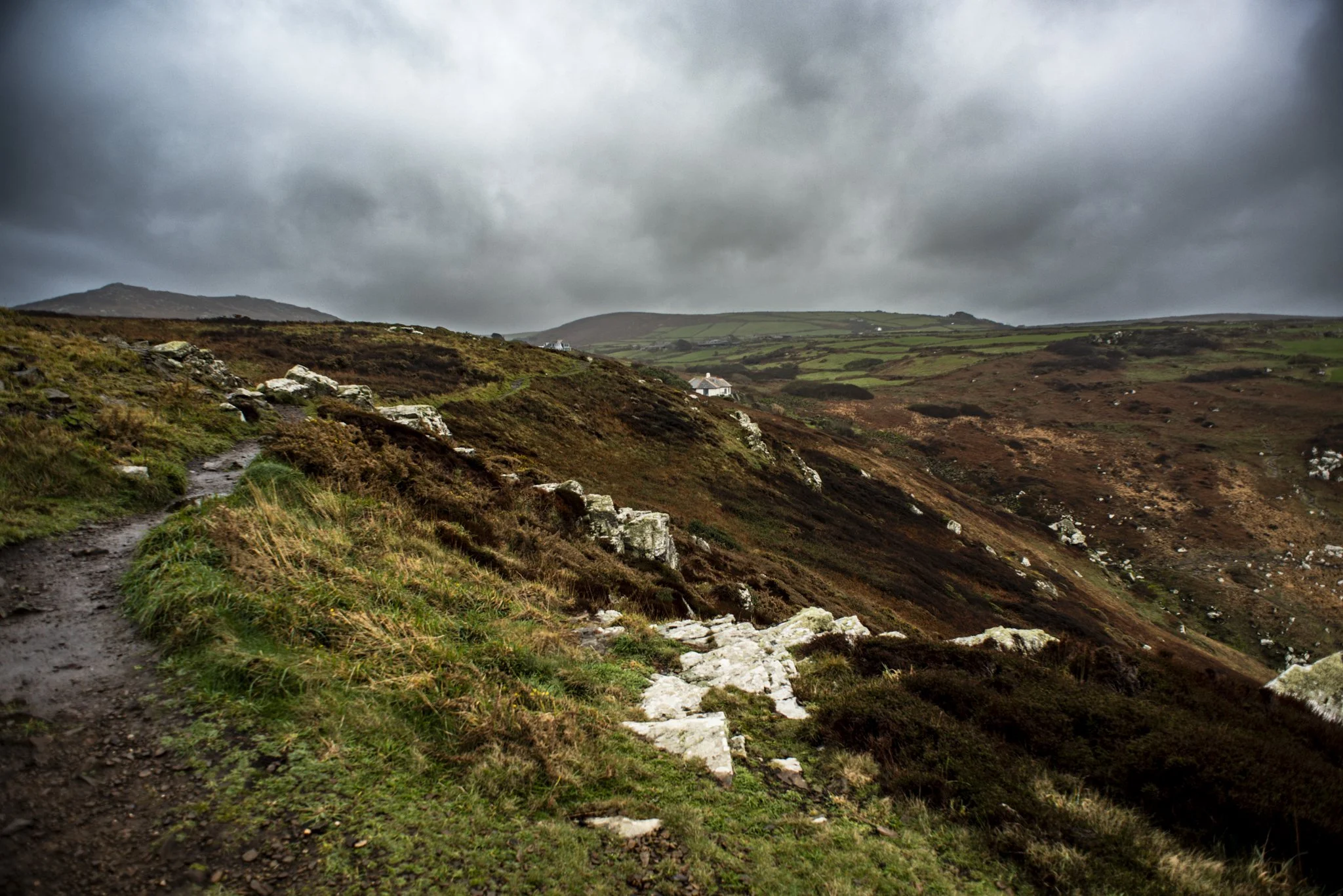 A rugged dirt trail winds along a grassy hillside with rocks scattered across the landscape, under overcast skies.