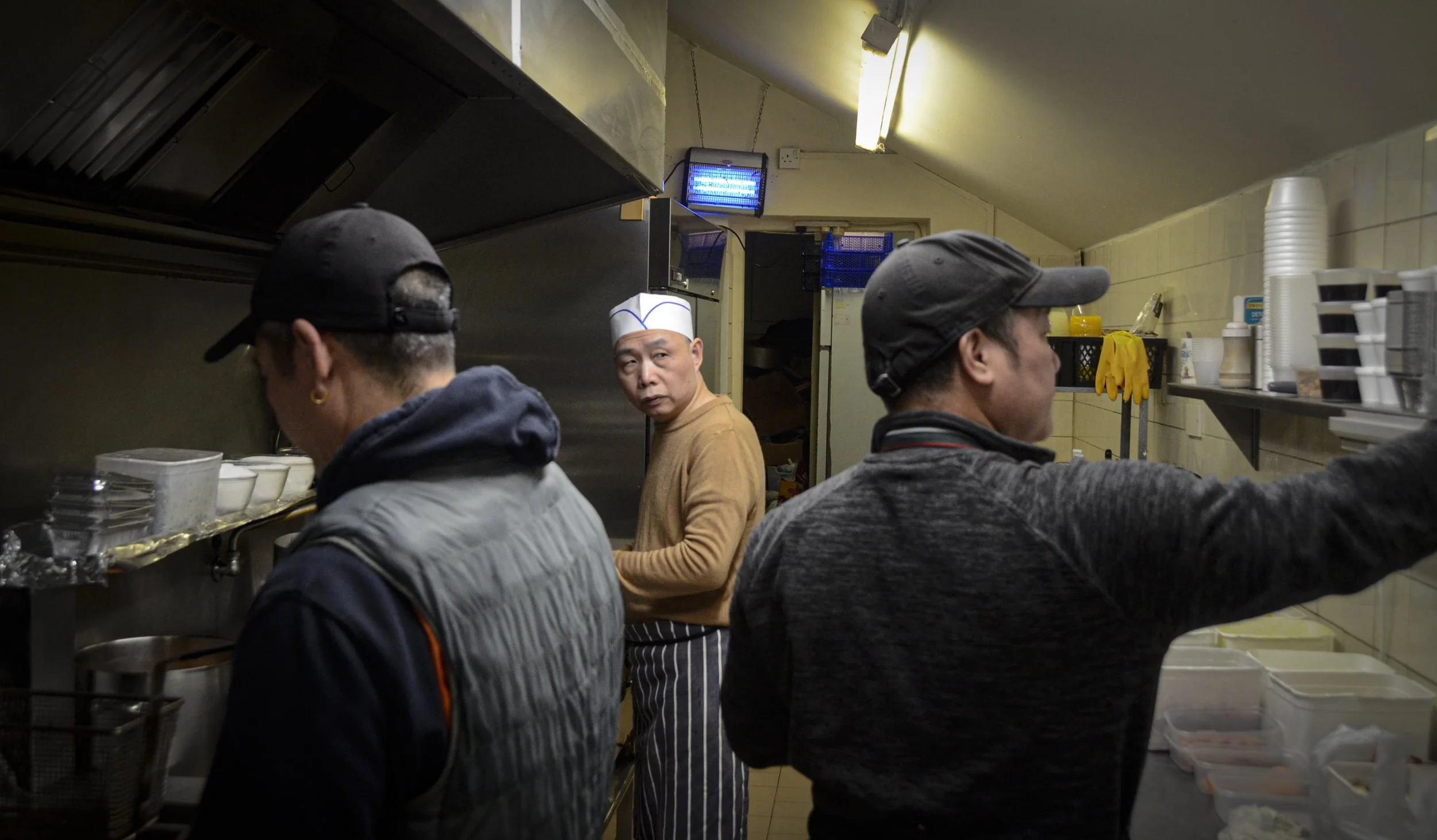 Three people working in a kitchen, one in the background wearing a white chef hat looking towards the camera, and two in the foreground wearing black caps and working with containers and utensils.