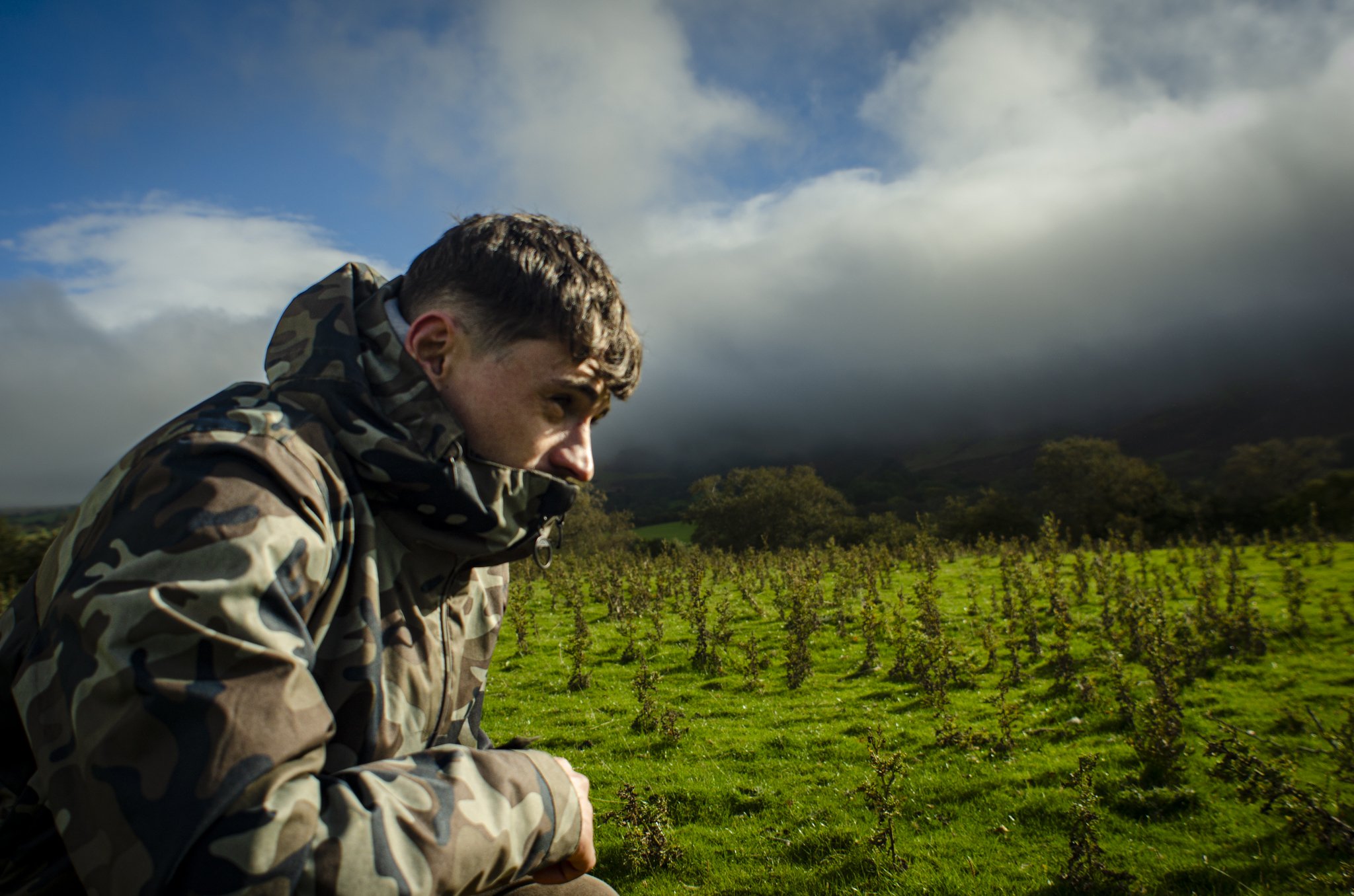 A man in camouflage jacket crouches in a green field with rows of plants, under a cloudy sky.