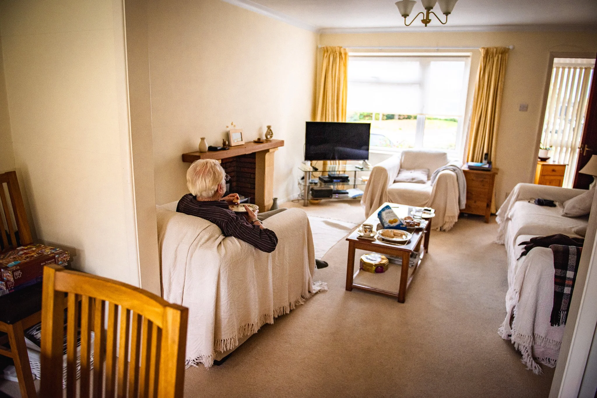 A living room with beige walls and carpet, featuring a woman with white hair sitting on a beige couch eating a meal, multiple beige sofas with throws, a wooden coffee table with food and drinks, a large window with yellow curtains, a flat-screen TV o
