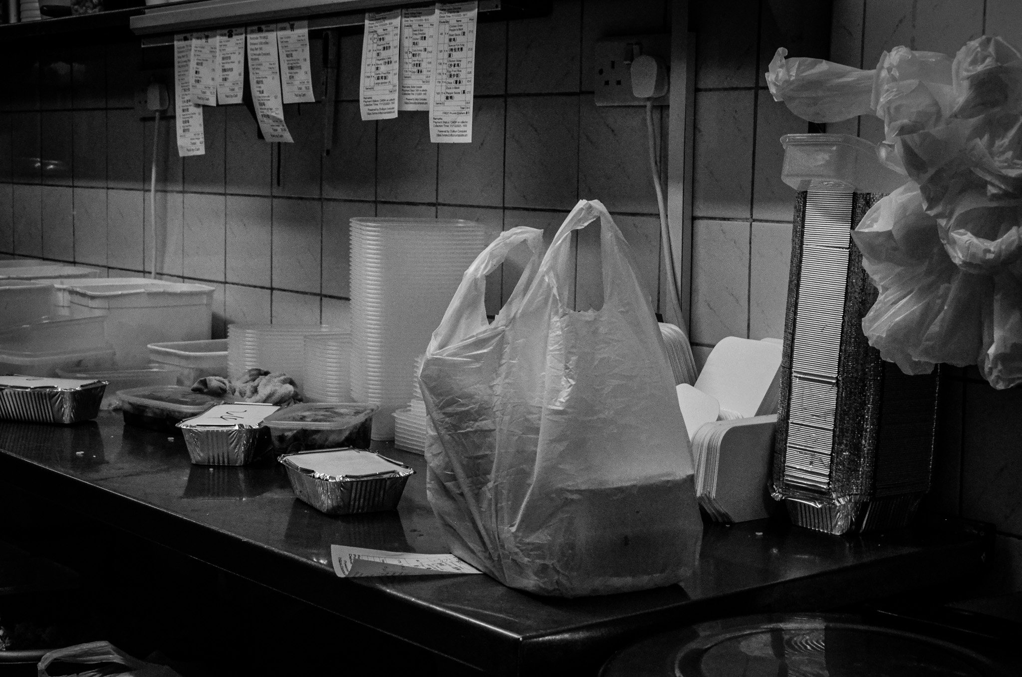 Takeout food containers, plastic bags, and disposable dishes on a kitchen counter with tiled wall.