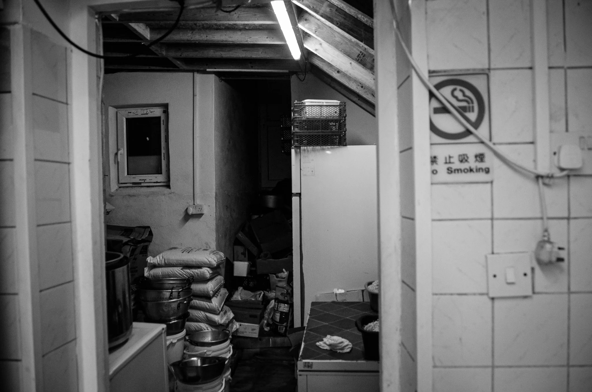 A black and white photo of a cluttered kitchen or storage room with stacked bags, bowls, and miscellaneous items, viewed through an open doorway.