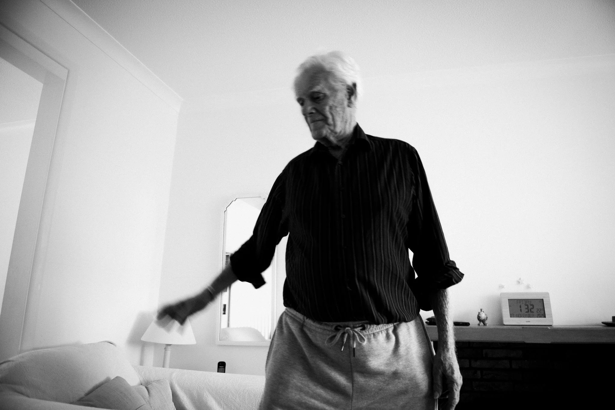An elderly man with white hair, wearing a dark, vertically striped shirt and light-colored sweatpants, standing and reaching towards a couch in a living room with a lamp, mirror, and clock visible in the background.
