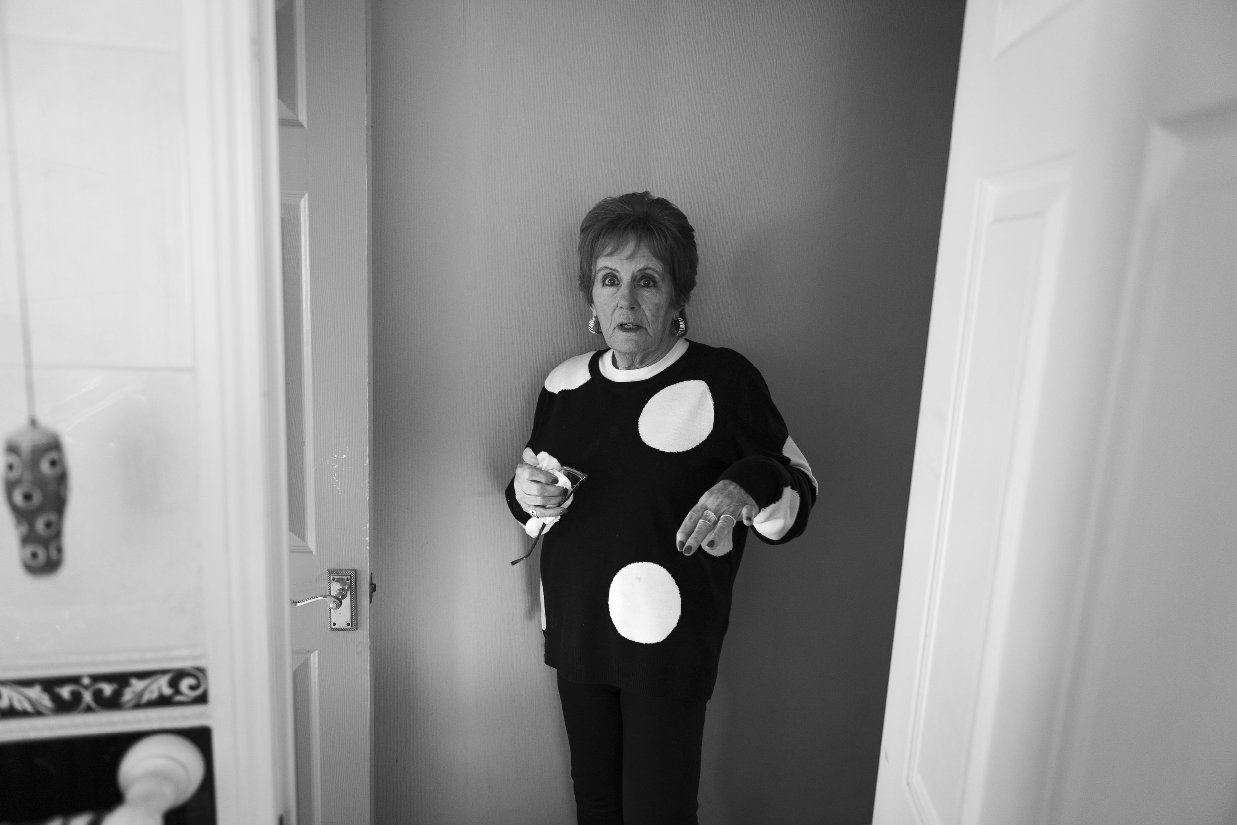 Older woman standing in doorway with surprised expression, wearing a sweater with large white polka dots.