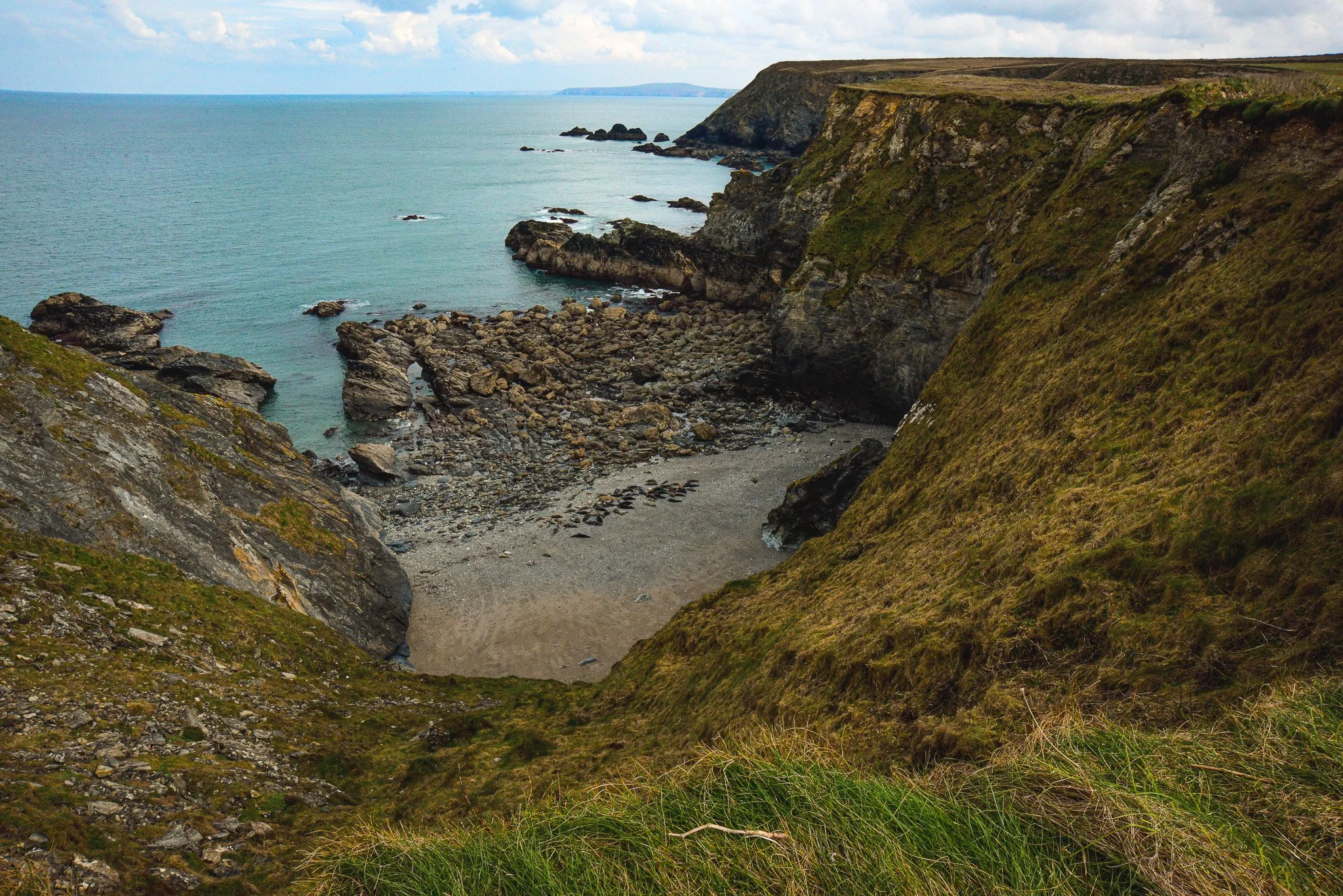 A view of a rocky beach with cliffs and green grass, overlooking the ocean with some clouds in the sky.