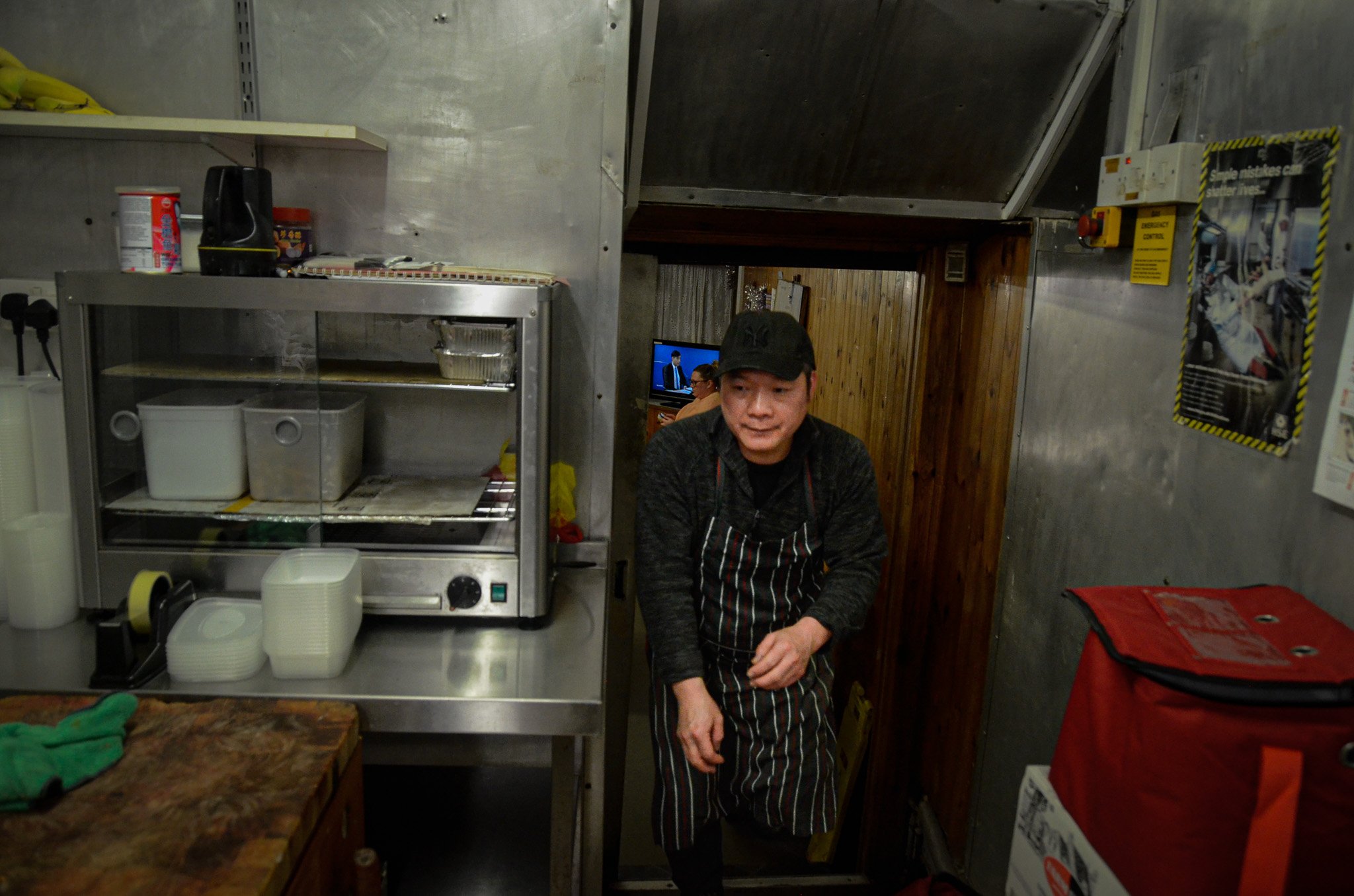 A man in a black cap and black apron with white stripes stands in a kitchen, in front of a metal food warmer and other kitchen supplies, with a small TV in the background.