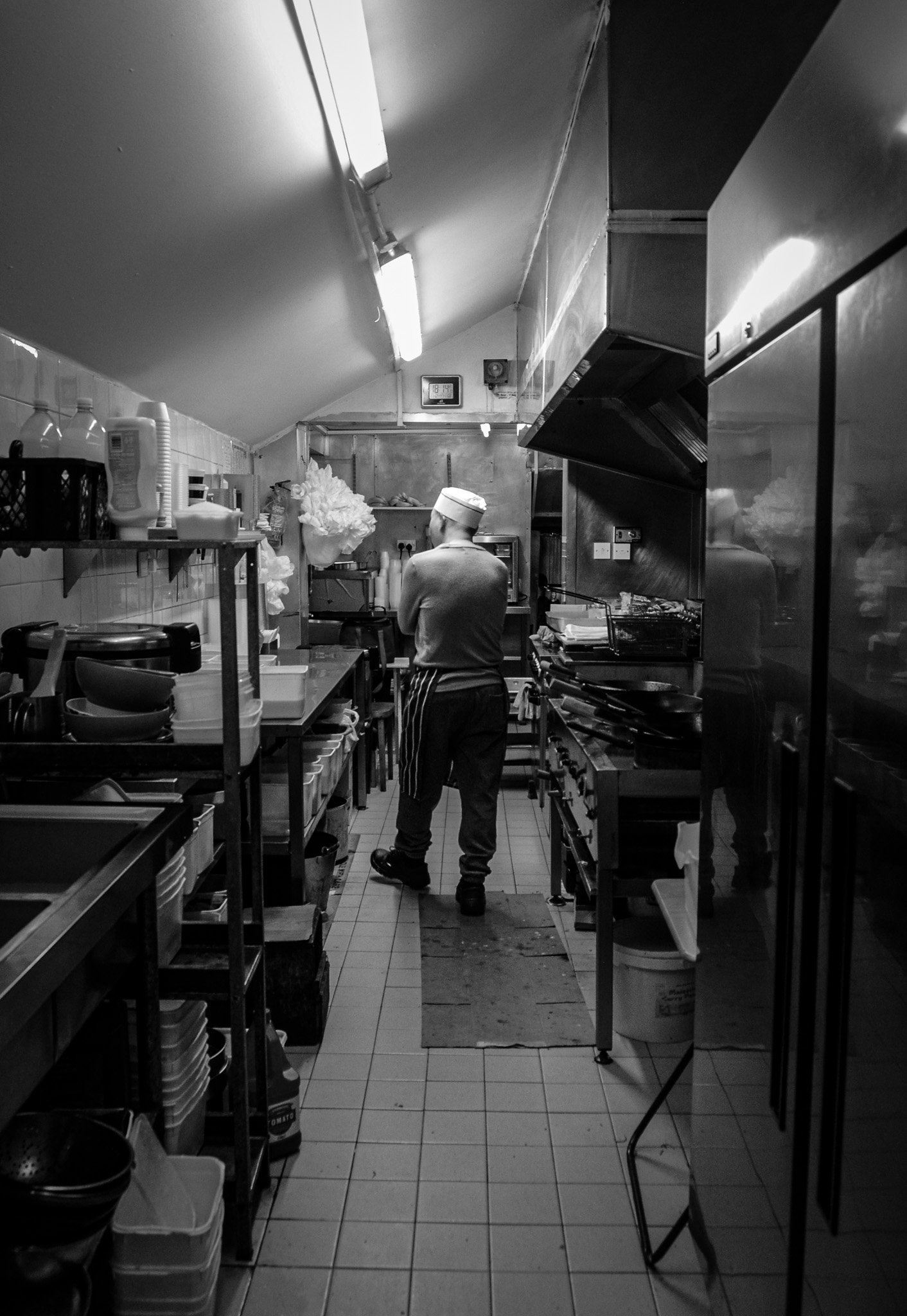 A chef in a commercial kitchen wearing a white hat and striped apron, working amid shelves with kitchen supplies, cooking appliances, and ingredients.