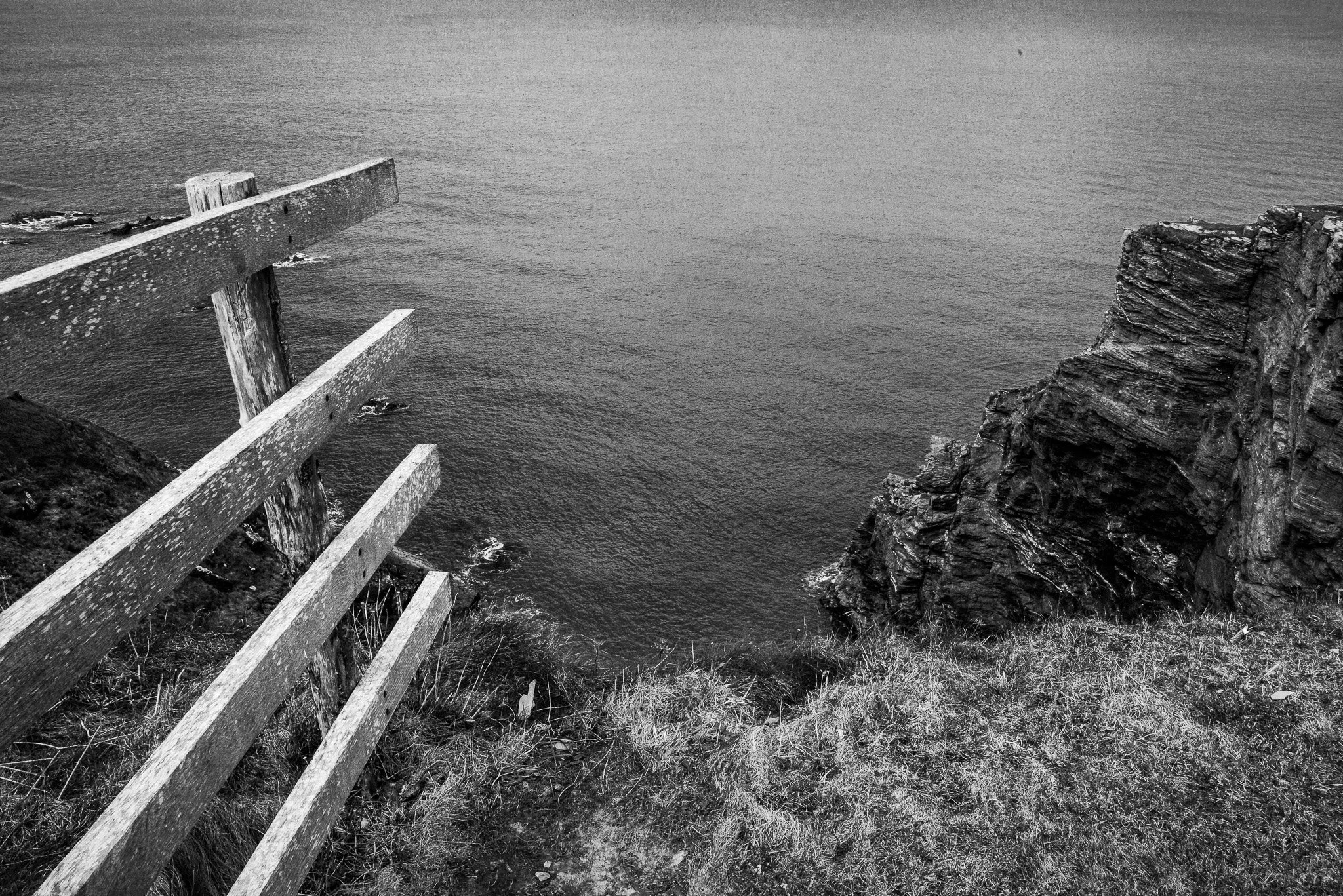Black and white photo of a wooden fence along a grassy cliff edge overlooking the ocean, with a large rock formation on the right.
