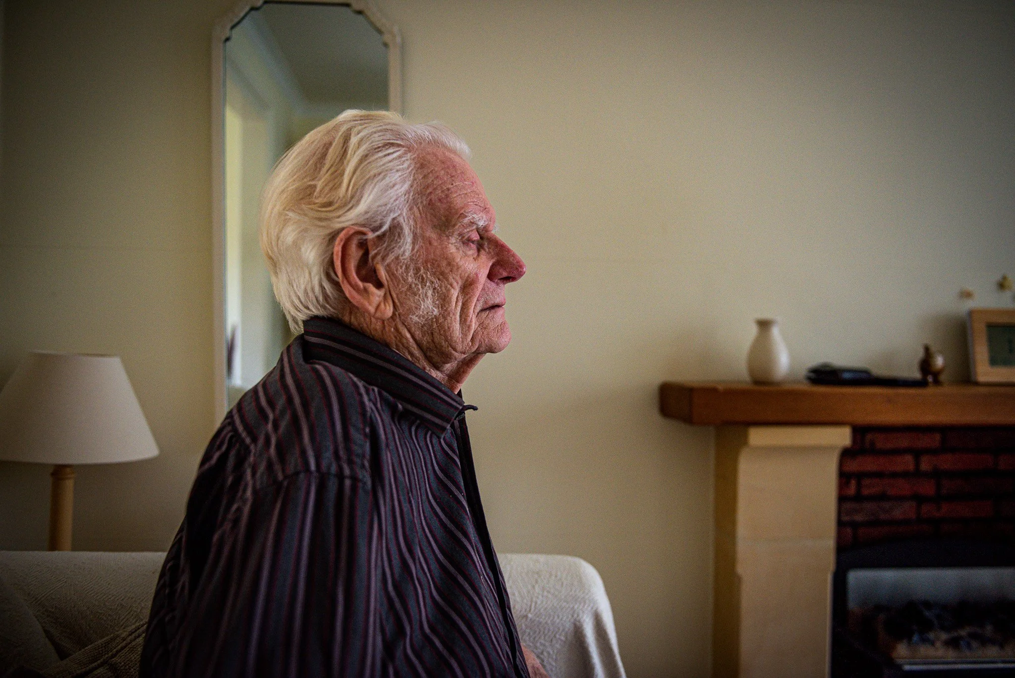An elderly man with white hair and a striped shirt sitting on a couch in a living room, with a fireplace and a mirror in the background.