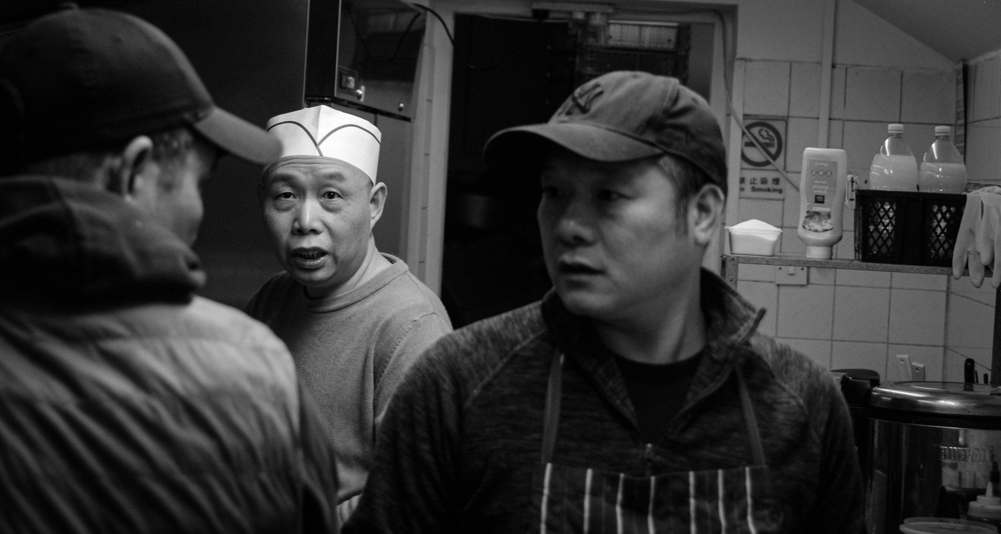 Three men in a kitchen, one wearing a paper chef's hat, another in a baseball cap, and the third in a cap, engaged in conversation.
