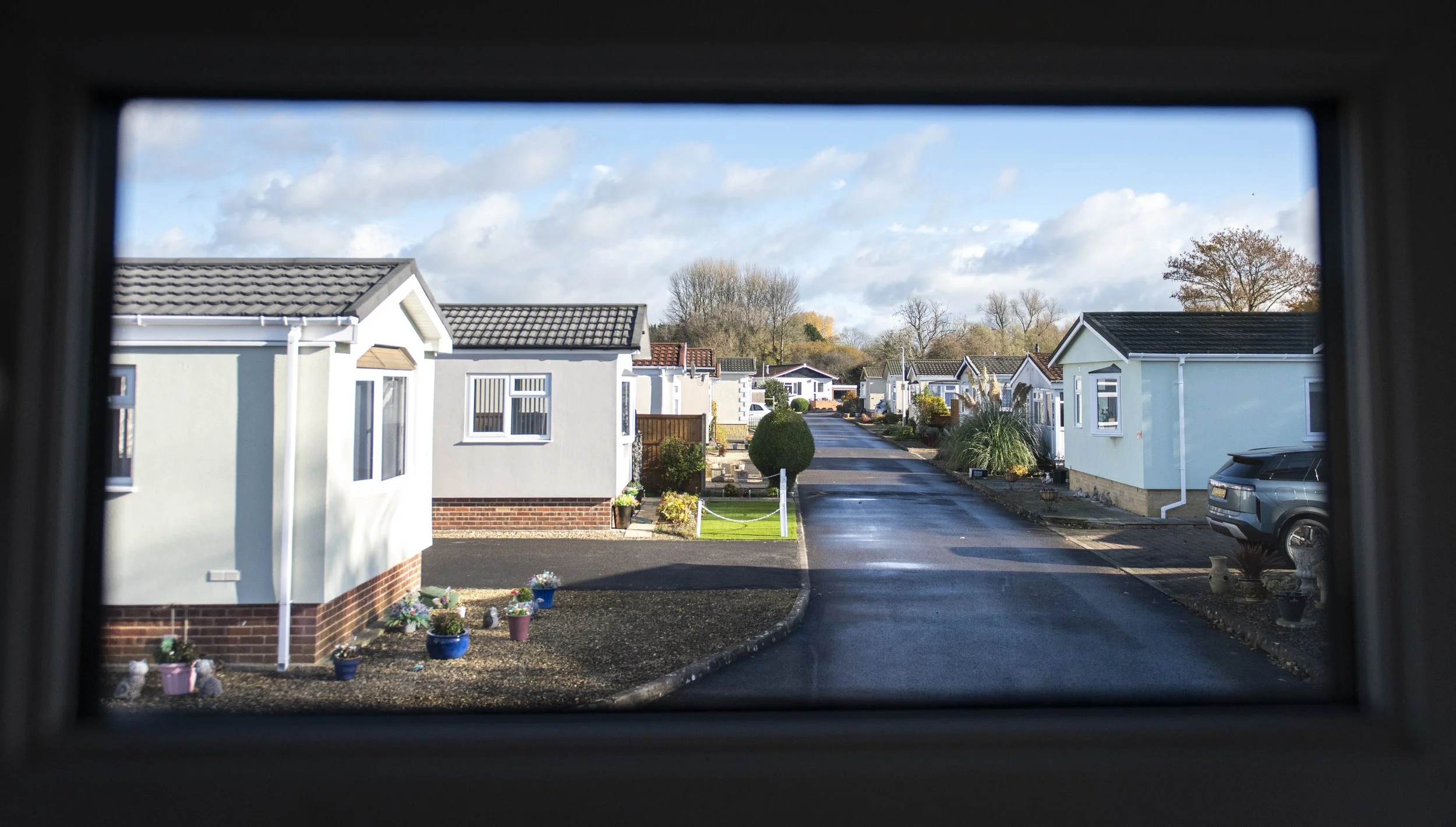 View of a suburban street seen through a window. Houses with manicured lawns and potted plants line both sides of the street under a partly cloudy sky.