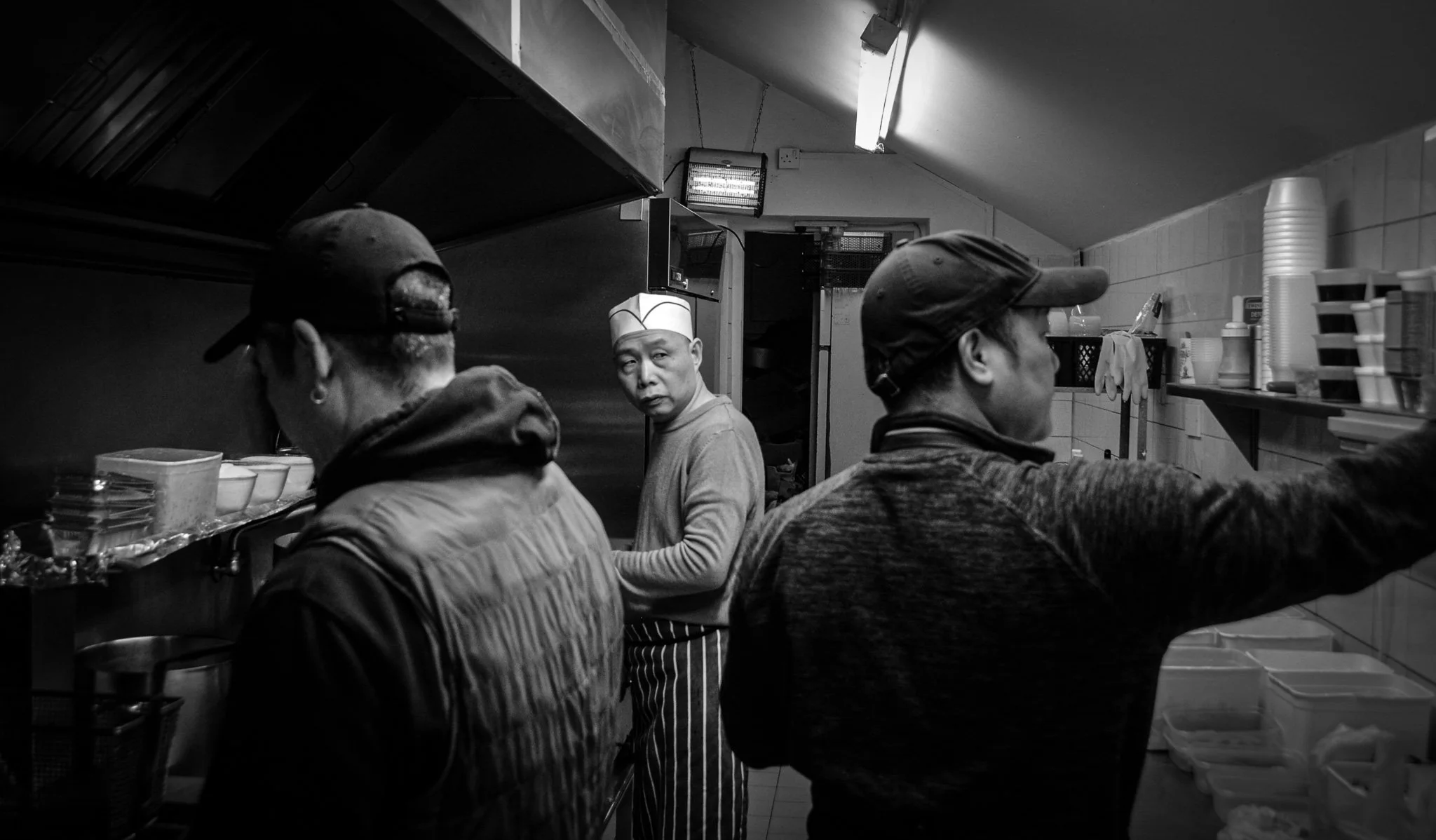 Black and white photo of three people working in a small kitchen, with one person looking directly at the camera. The person in the center wears a paper hat and striped apron, while the others wear caps and casual clothing, surrounded by kitchen supp