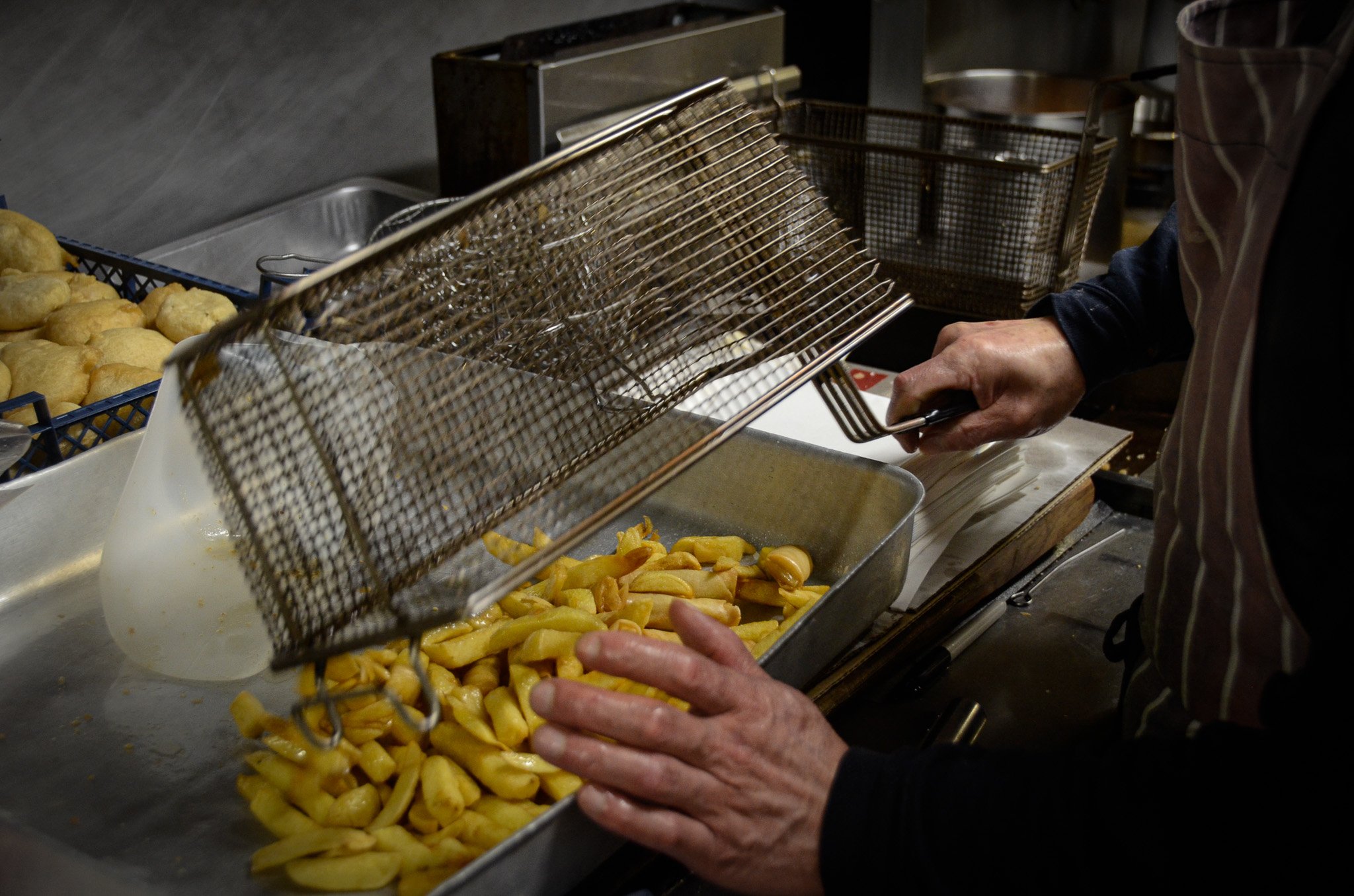 Person scooping fried yellow potato wedges from a metal tray into a paper bag in a kitchen or restaurant setting.