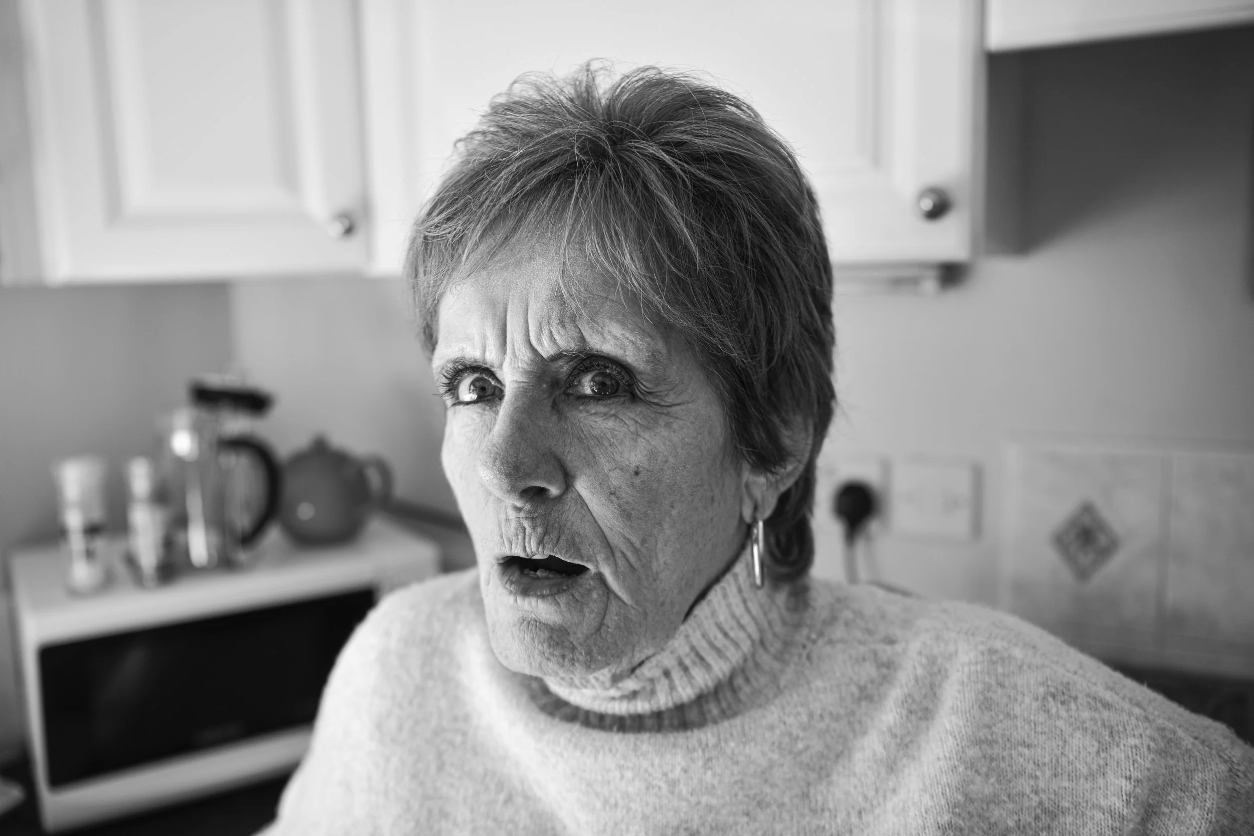 Black and white photo of an elderly woman with short hair, wearing a turtleneck sweater, with a surprised or confused expression, in a kitchen setting.