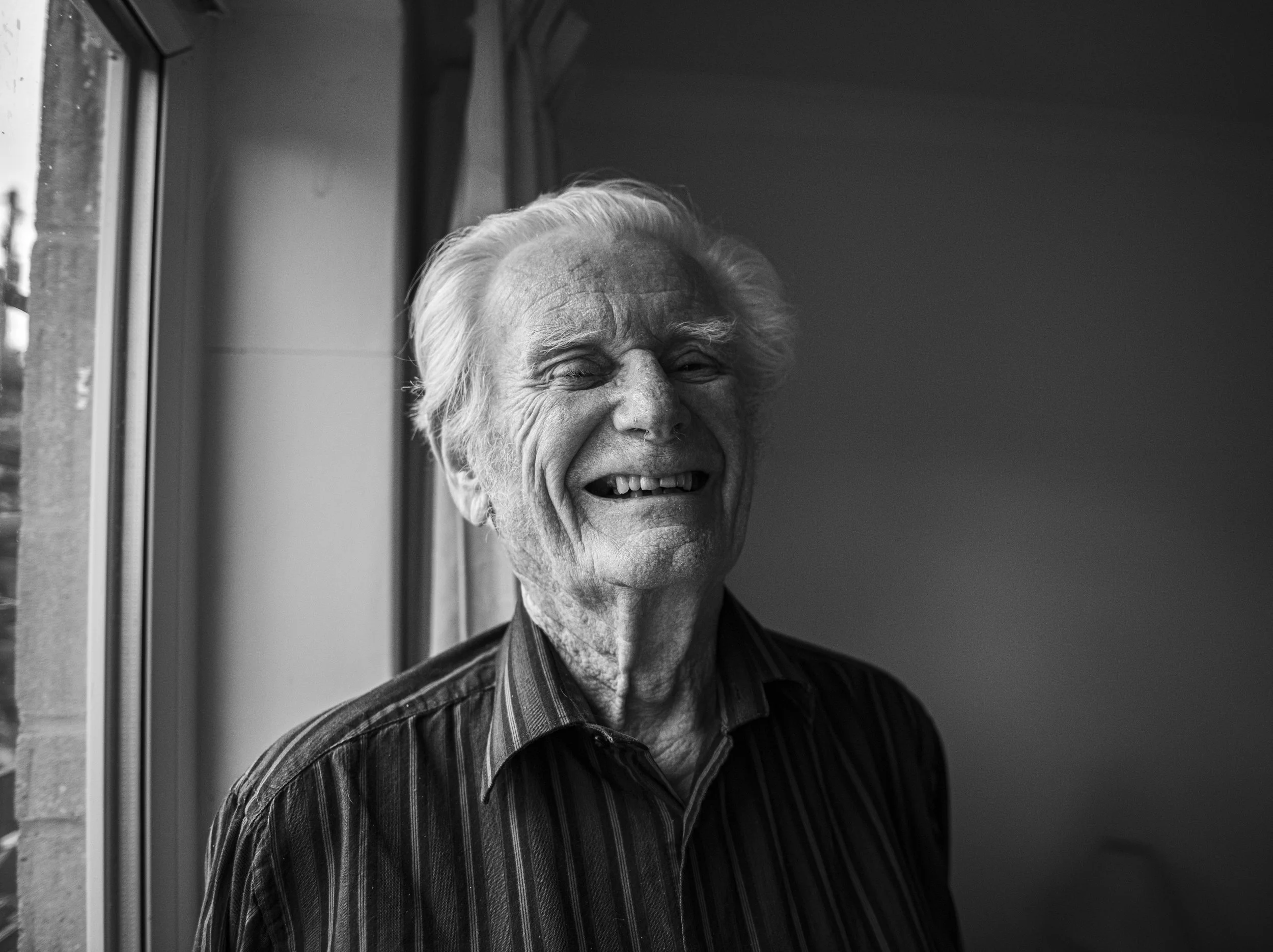 A black and white photo of an elderly man smiling with missing teeth, standing by a window with curtains, wearing a striped shirt.