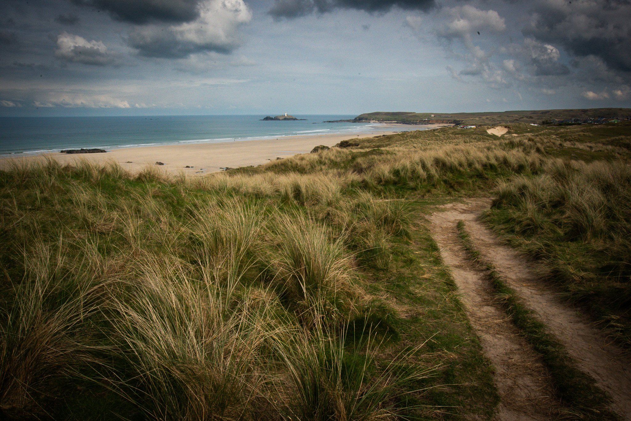 Dirt path winding through grassy dunes leading to a sandy beach with ocean and lighthouse in the distance under dark cloudy sky.