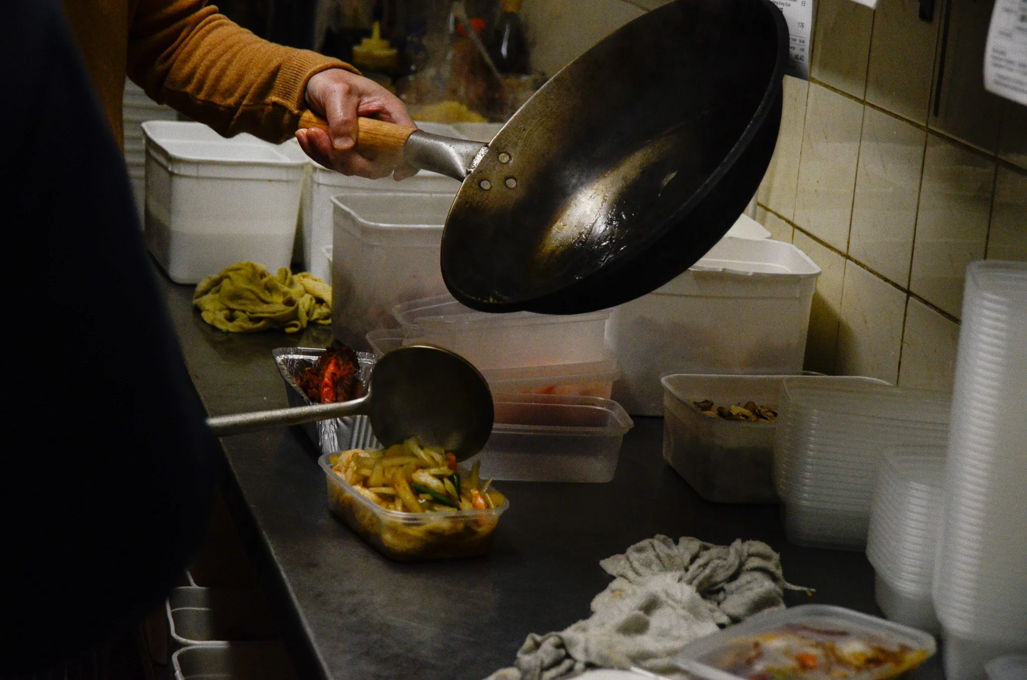 Person pouring stir-fried vegetables from a wok into a container at a food service counter.