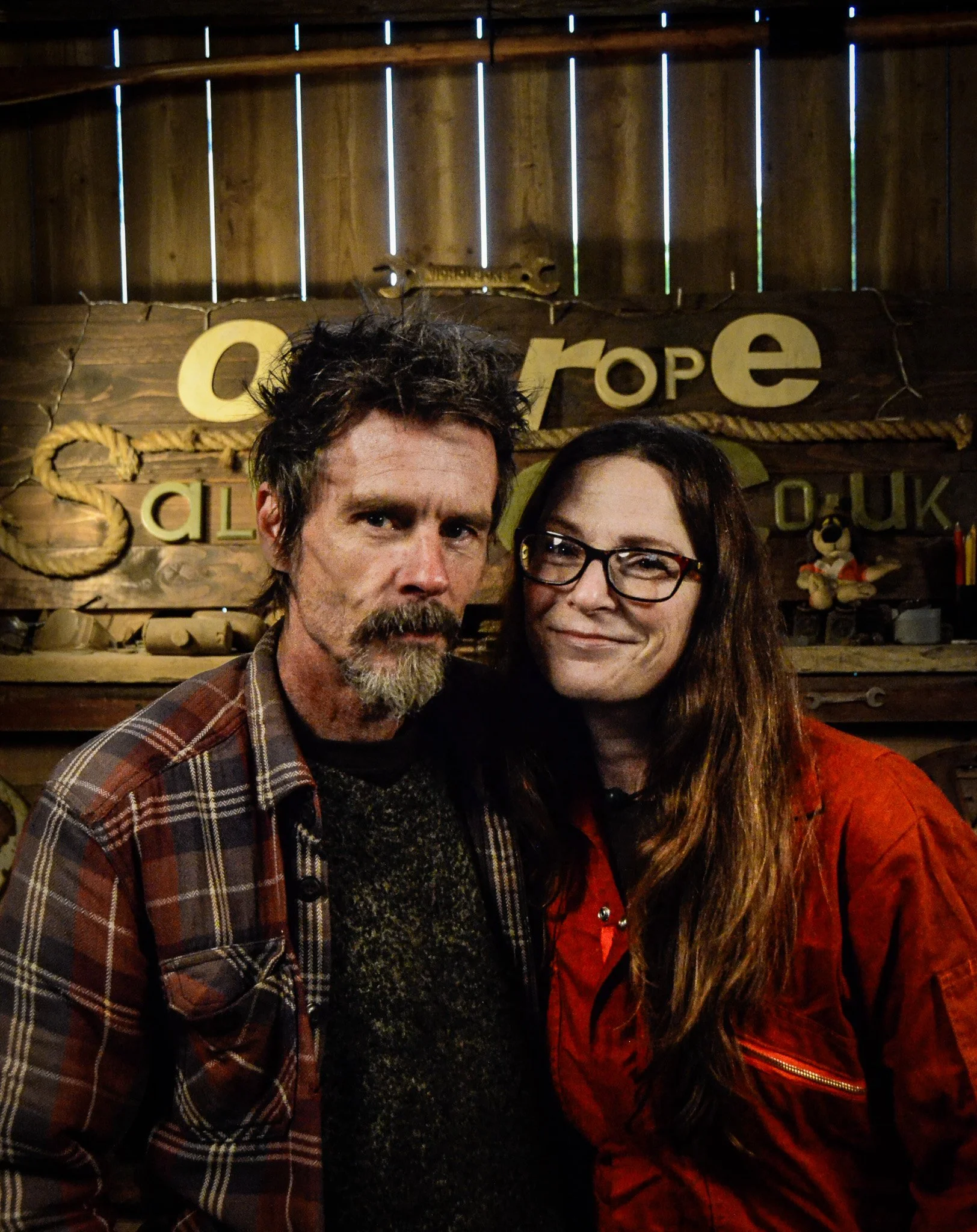 Two people posing together in front of a wooden wall with a sign that reads 'Qu Erope'. One person has a beard, plaid shirt, and dark curly hair, while the other has long brown hair, glasses, and a red jacket.