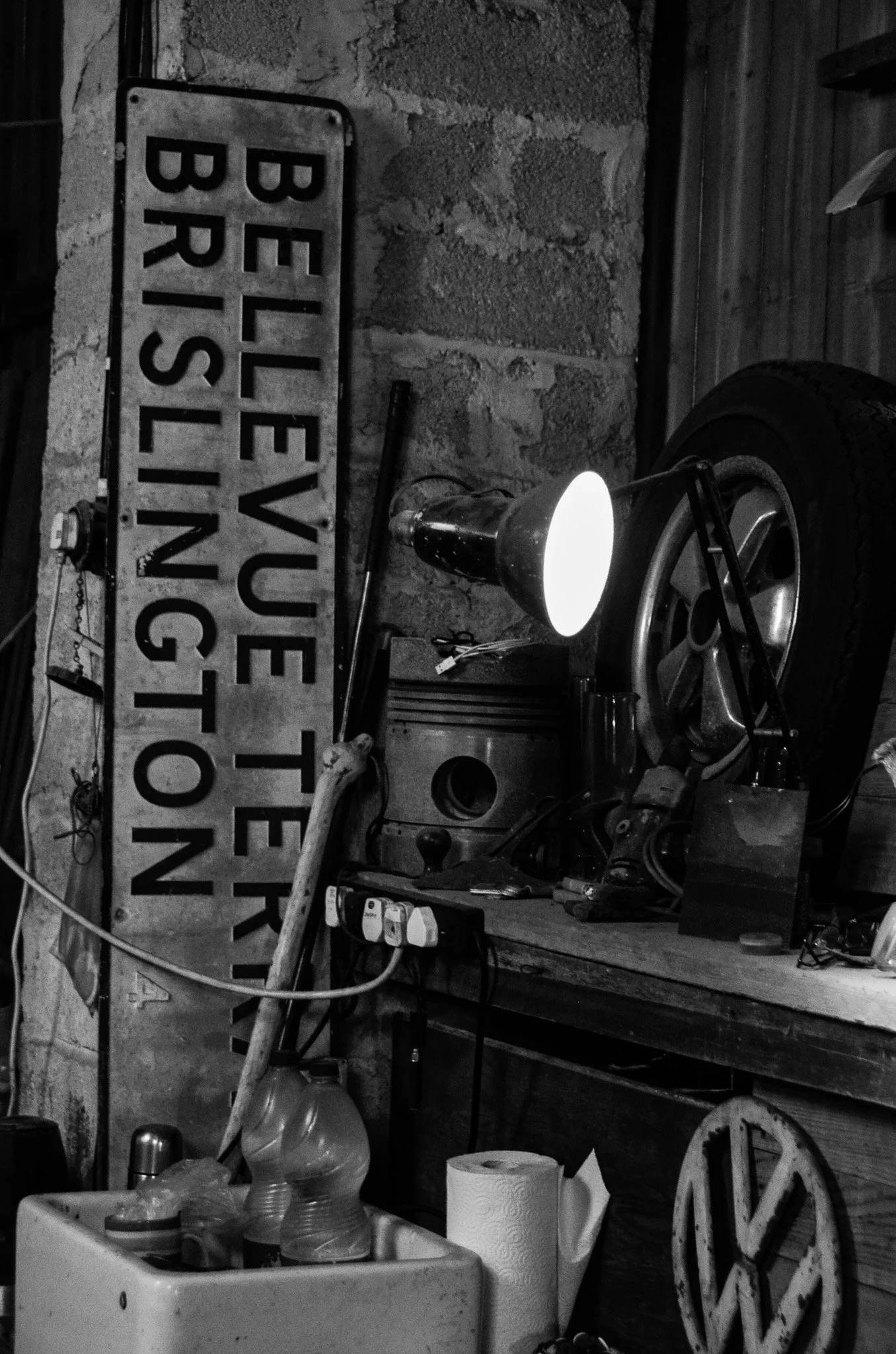 Black and white photo of a cluttered garage or workshop with a vintage 'BELLEVUE TRAINING' sign, a car wheel, and various tools and objects on a wooden workbench.