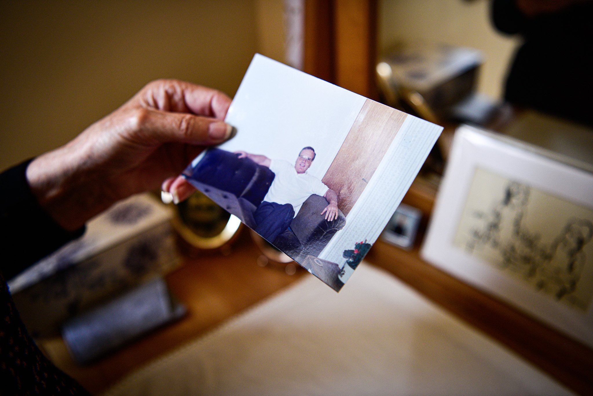 Hand holding a printed photo of a man sitting on a brown armchair in a room with wood paneling and framed artwork on the wall.