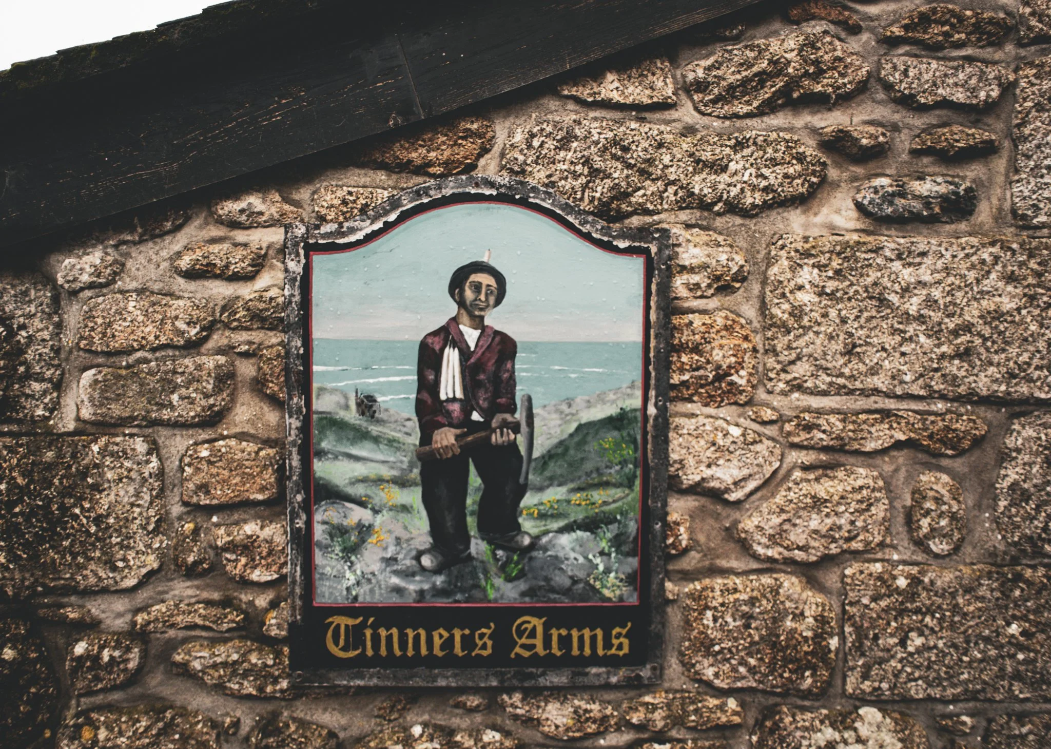 A painted sign on a brick wall depicting a historical figure working with a pickaxe, with a landscape background, and the words "Tinners Arms" written at the bottom.