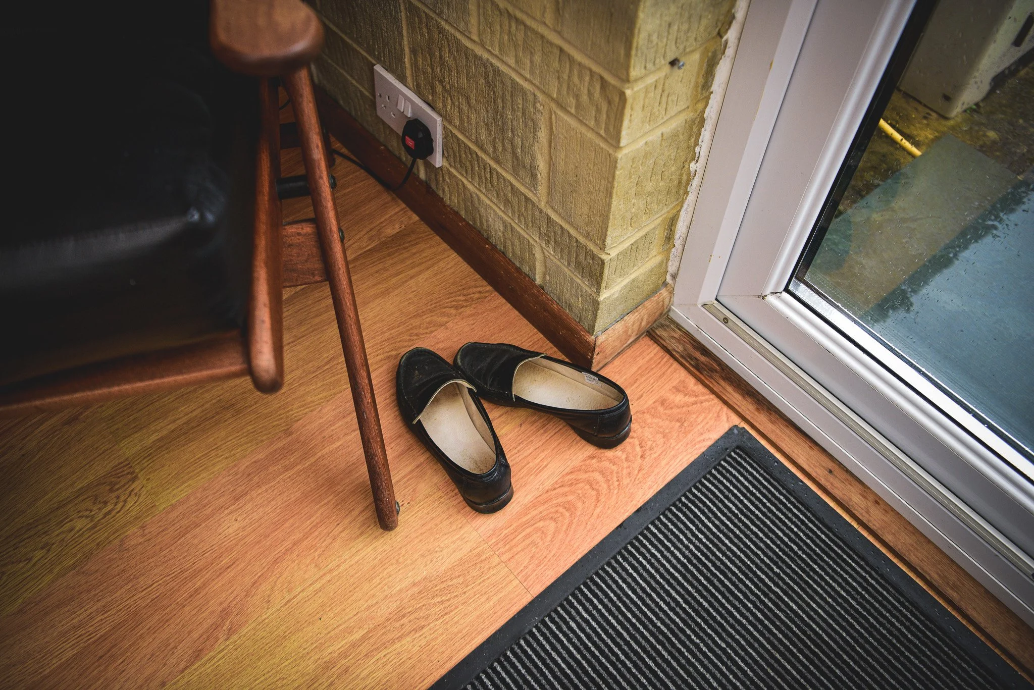 A pair of black shoes on a wooden floor near a glass door. There is a chair with wooden legs and a black shoe on it, and a brick wall with an electrical outlet nearby.