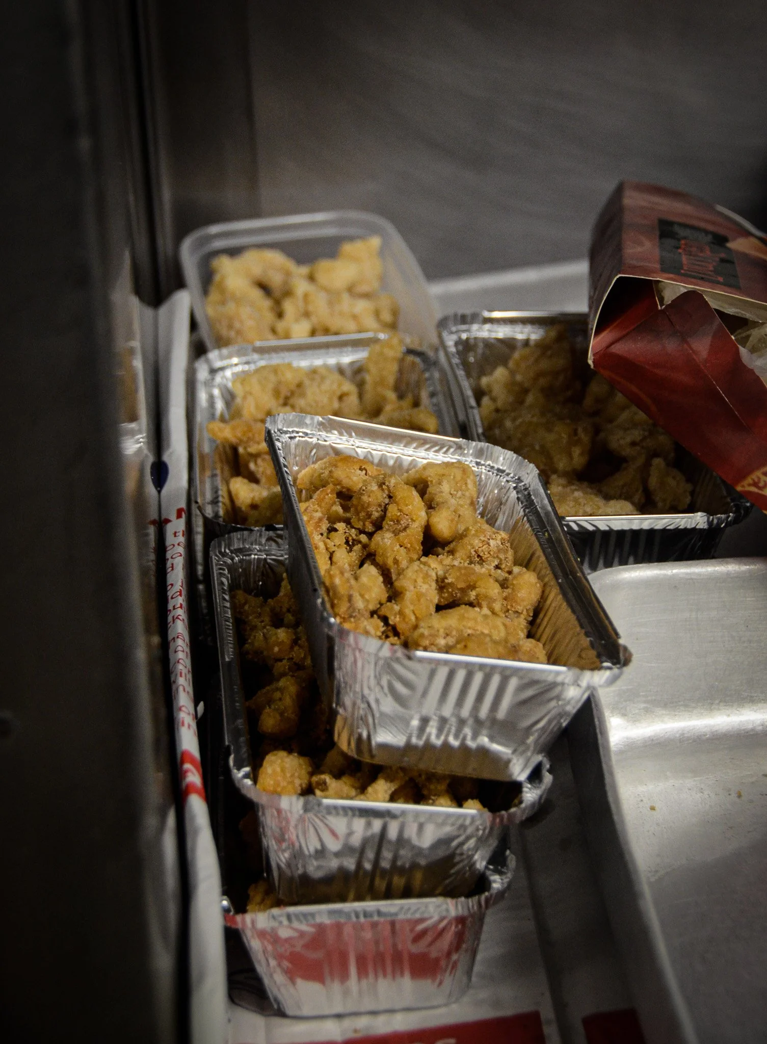 Several aluminum containers filled with fried chicken pieces, placed inside a fast food restaurant on a counter near a silver tray.
