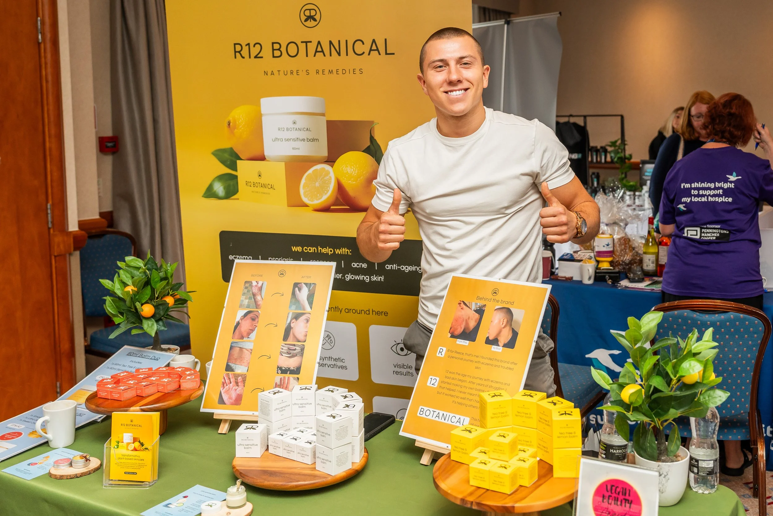 A man smiling at a booth promoting R12 Botanical skincare products, with various skincare products on display, informational posters, and potted plants around.