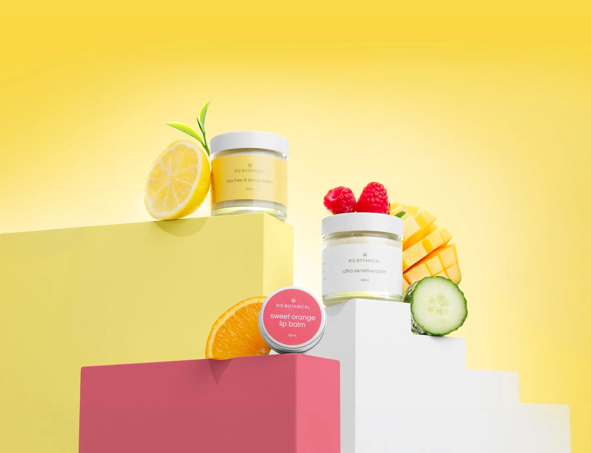 A collection of skincare products, including R12 Botanical tea tree & lemon balm balm, ultra sensitive balm, and sweet orange lip balm, surrounded by slices of lemon, raspberry, cucumber, orange, and mango against a yellow and pink background.