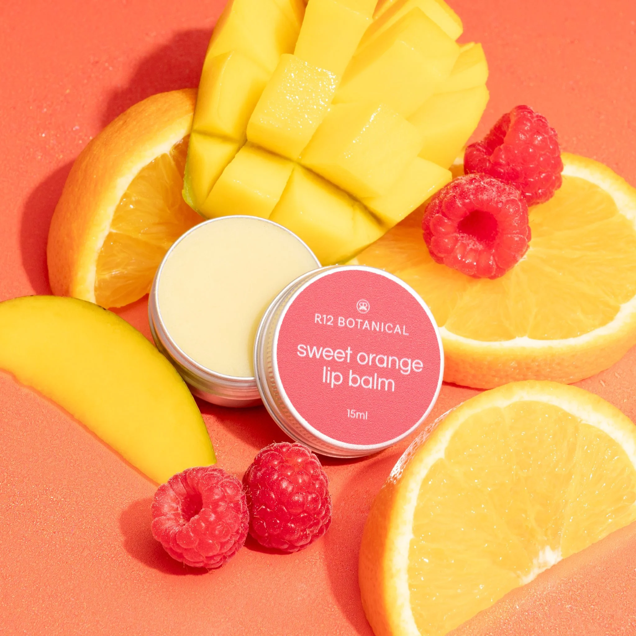 Close-up of sliced orange, yellow mango, and raspberries on bright orange background, with a container of lip balm labeled 'R12 Botanical Sweet Orange Lip Balm' in the center.
