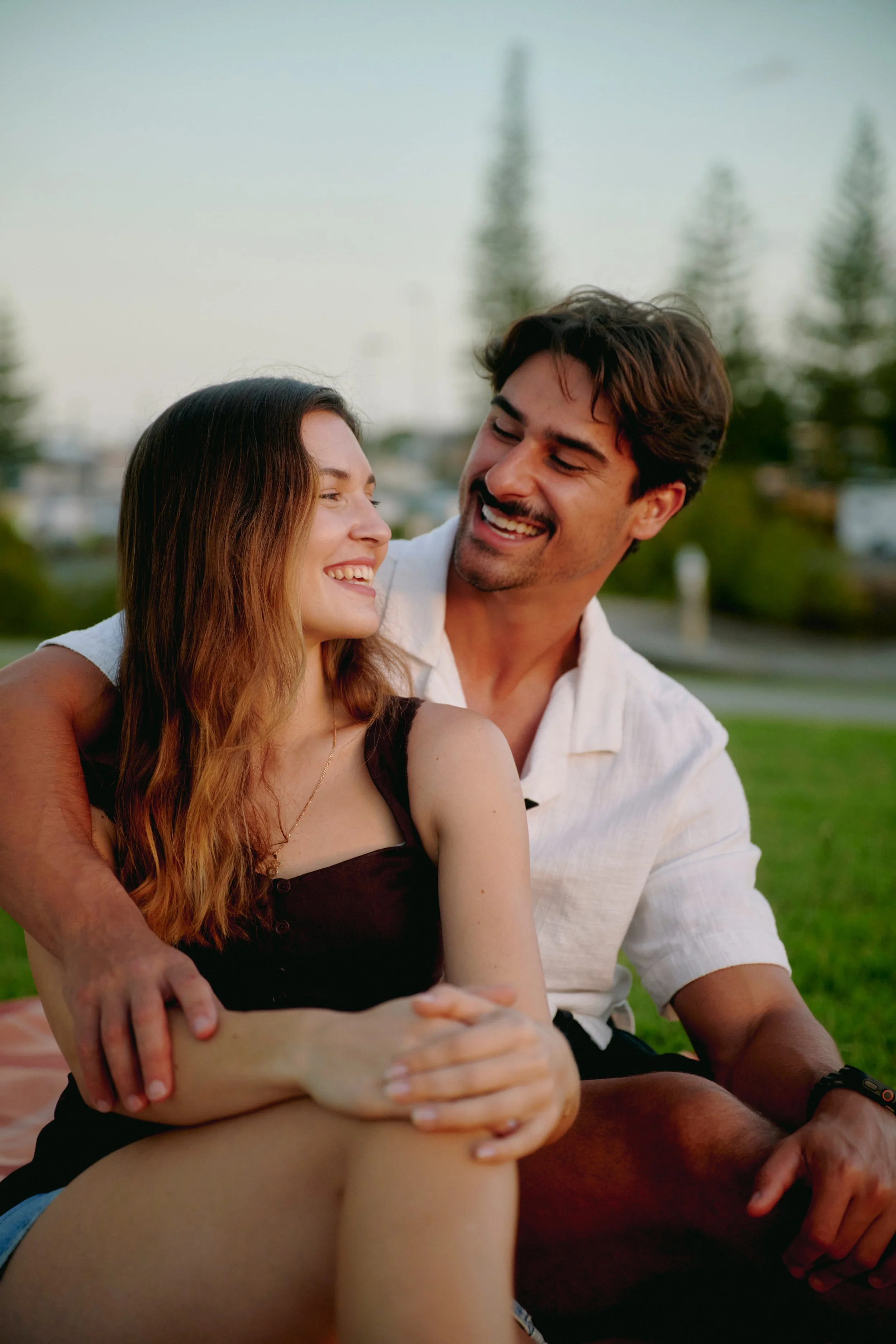A smiling woman with long brown hair sitting on a man's lap in a park with trees in the background.