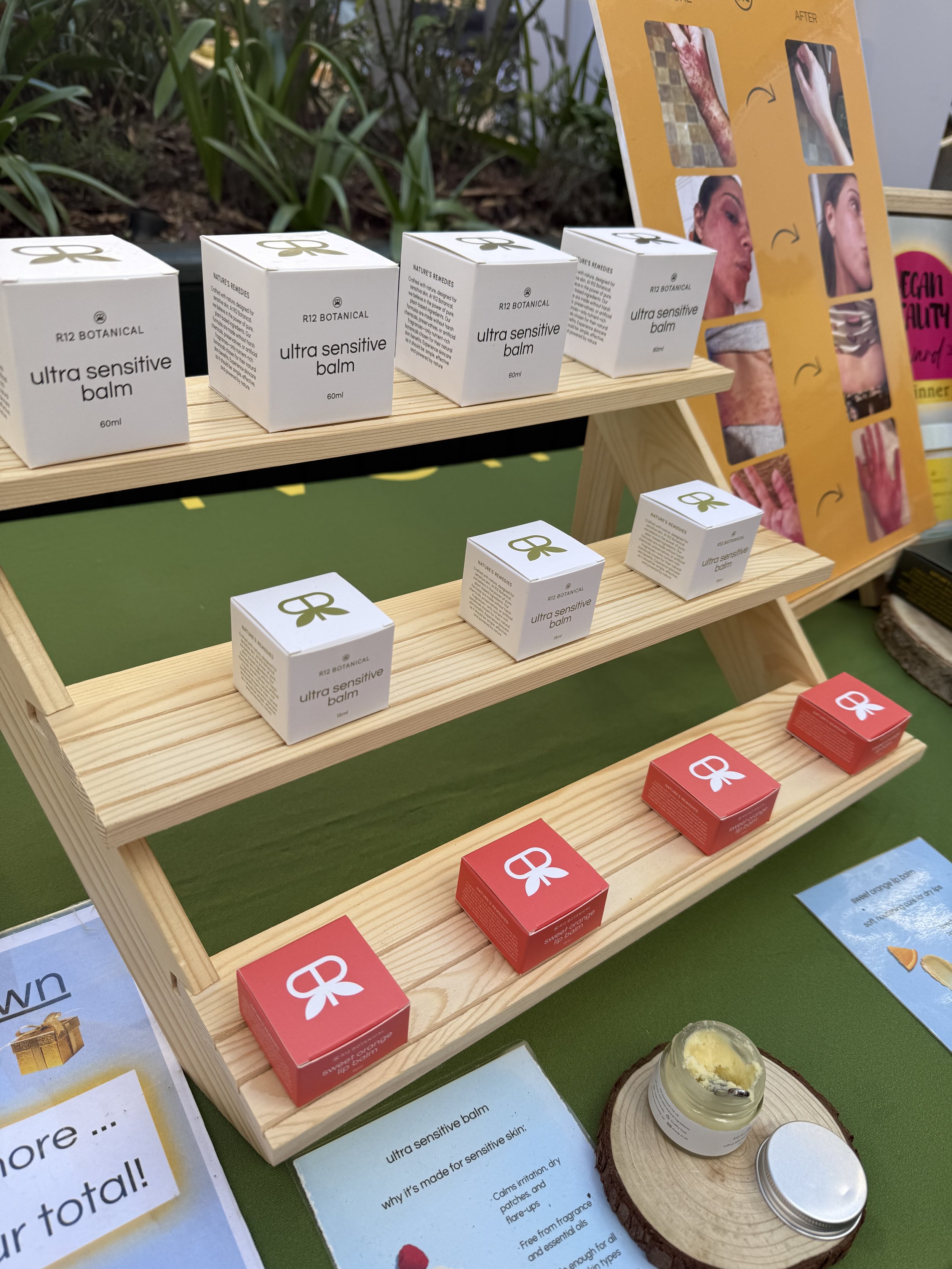 Display of skincare products, specifically ultra sensitive balm, arranged on a wooden shelf. The products are in white and red boxes with a bow logo. Behind the shelf is a yellow informative poster with images showing before and after skin treatments. There is also a small jar of balm on a wooden coaster at the bottom right of the image.