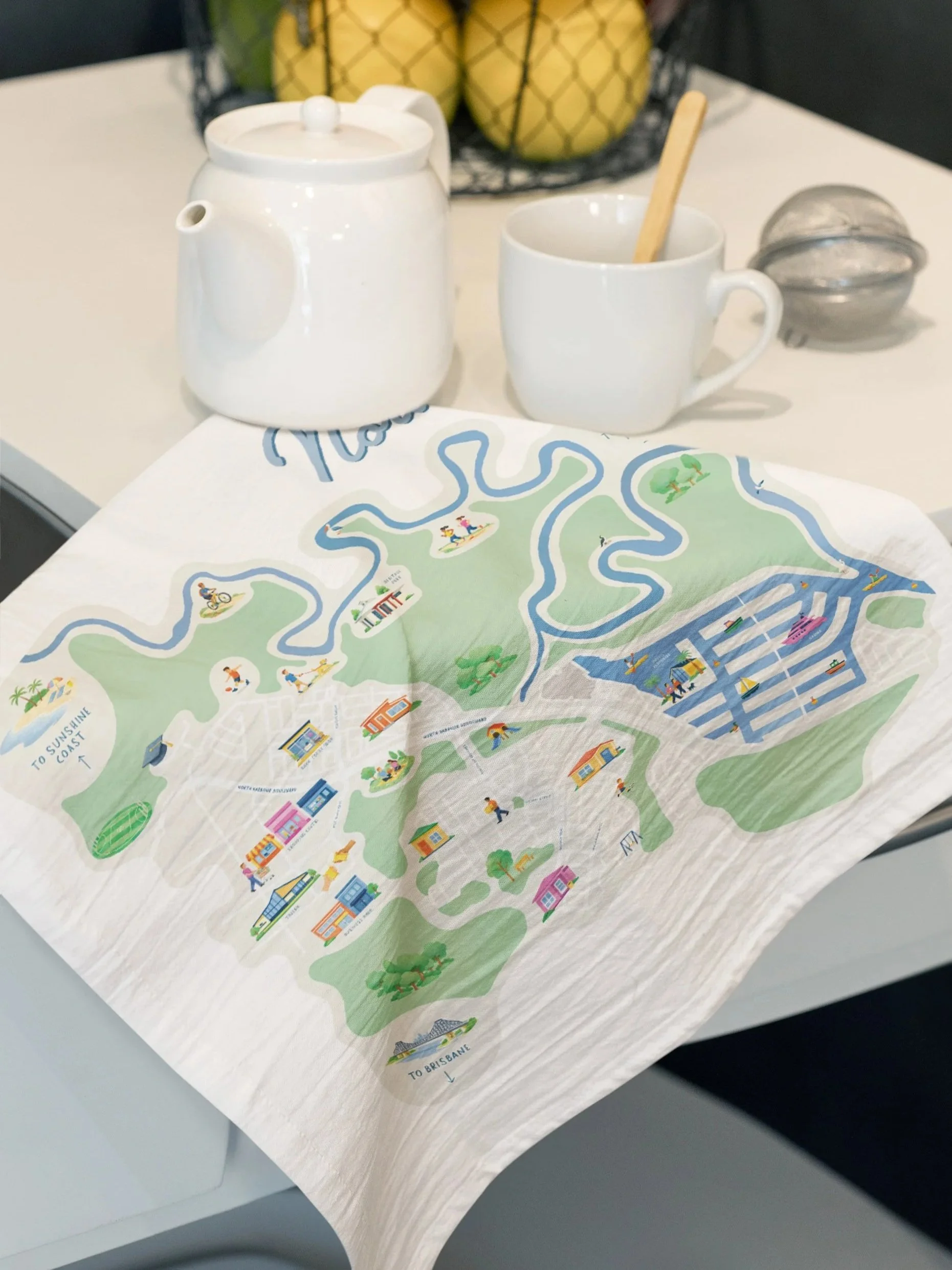 Colorful illustrated map showing a river, parks, buildings, and pathways with nearby items like a white ceramic teapot, a cup with a wooden stirrer, a metal container, and a bowl of fruit in the background on a table.