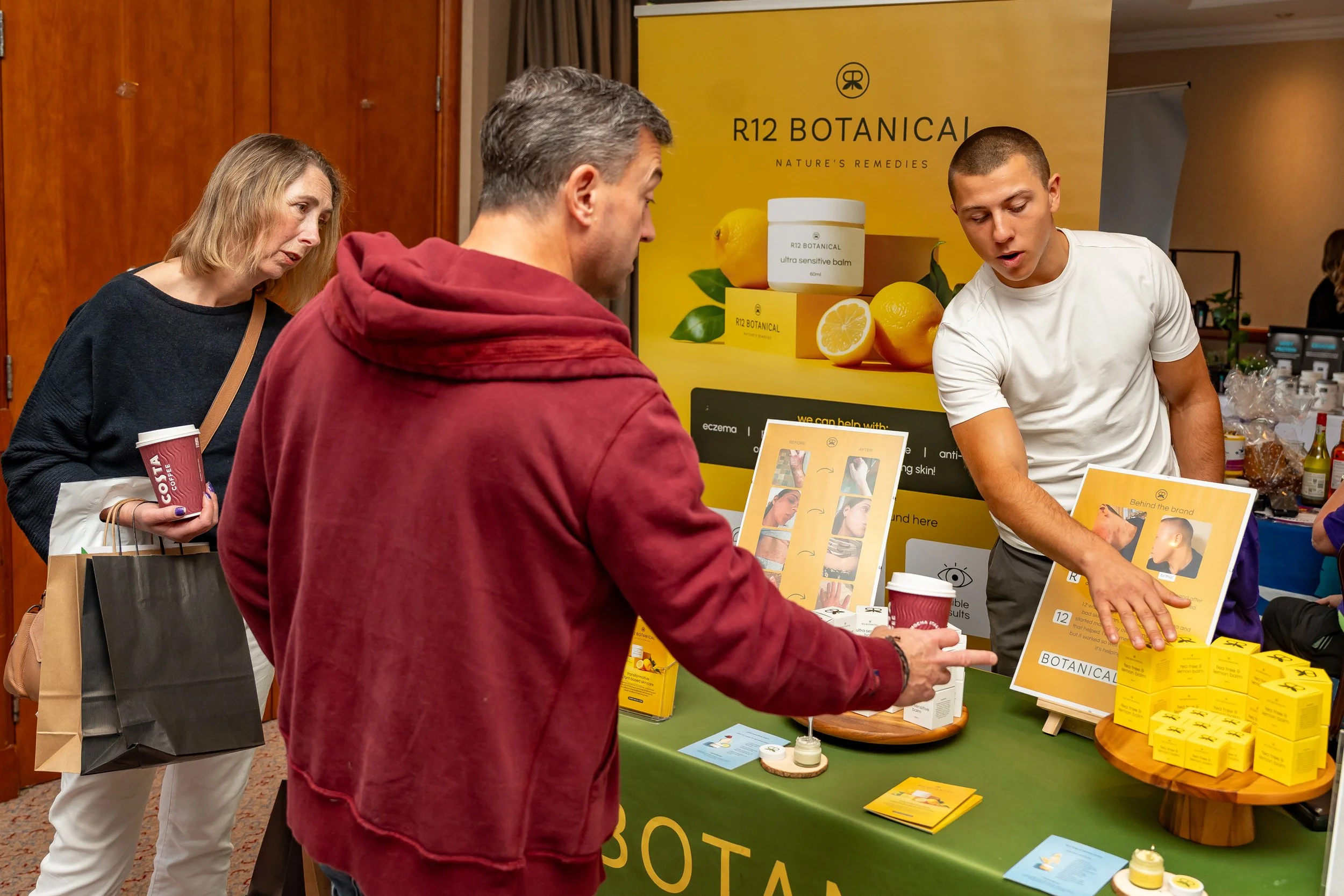 People are at a booth displaying R12 Botanical skincare products. A man in a red hoodie is looking at the products, another woman with long blonde hair and a black shirt is holding a Costa coffee cup and a shopping bag, and an employee in a white shirt is explaining the products.