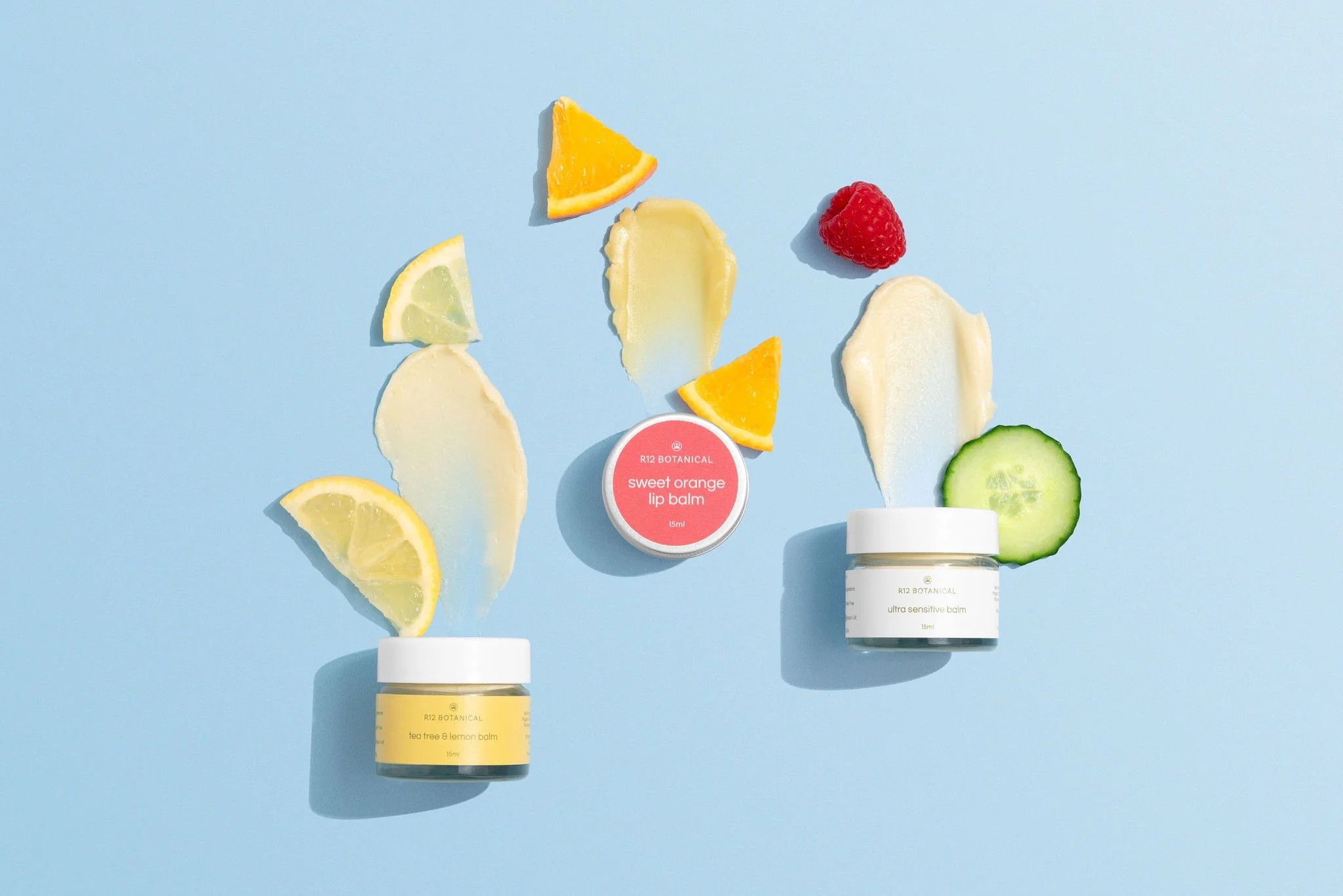 Three small jars of lip balm with sliced lemon, orange, cucumber, and raspberry placed around them on a light blue background.