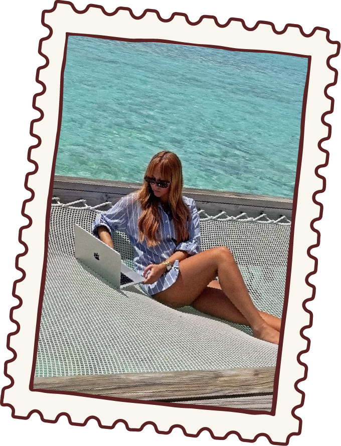 A woman with long red hair and sunglasses relaxing on a hammock by the water, working on a laptop.