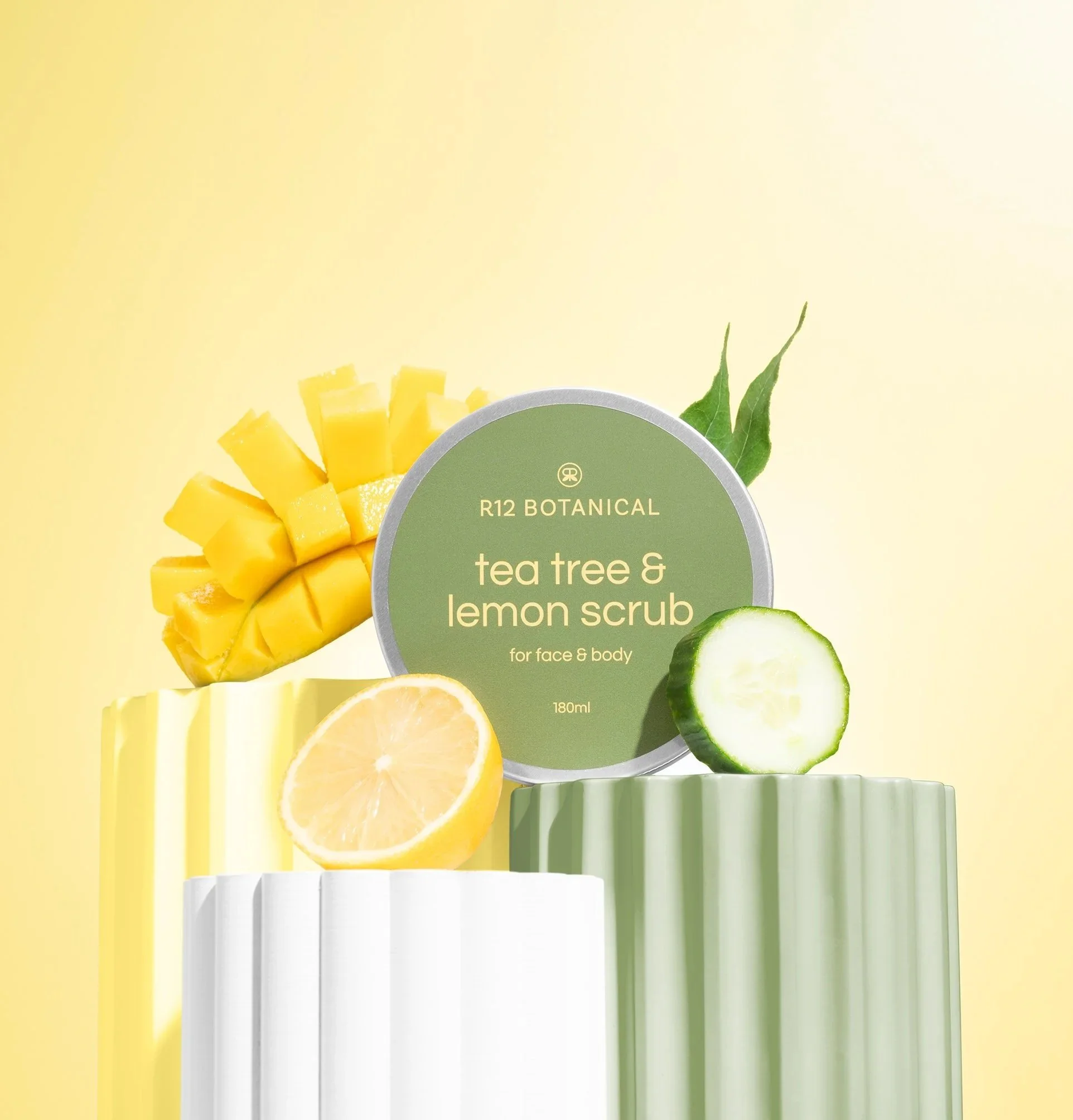 A cosmetic product labeled "R12 Botanical Tea Tree & Lemon Scrub" with sliced lemon, mango, cucumber, and green leaves around it, on a yellow background.