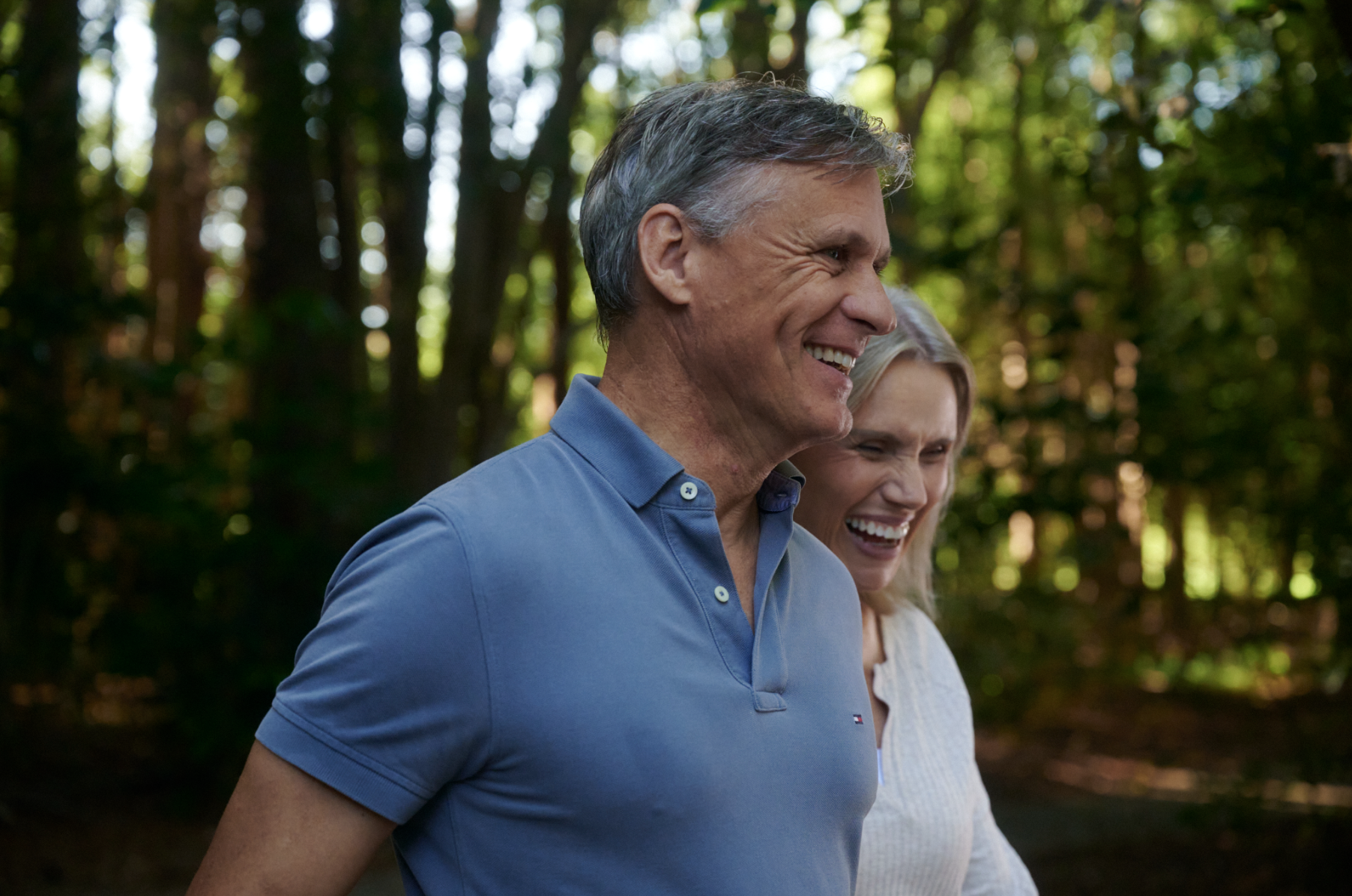 A smiling middle-aged man in a blue polo shirt walking outdoors with a woman in a white sweater, with a background of trees and sunlight.