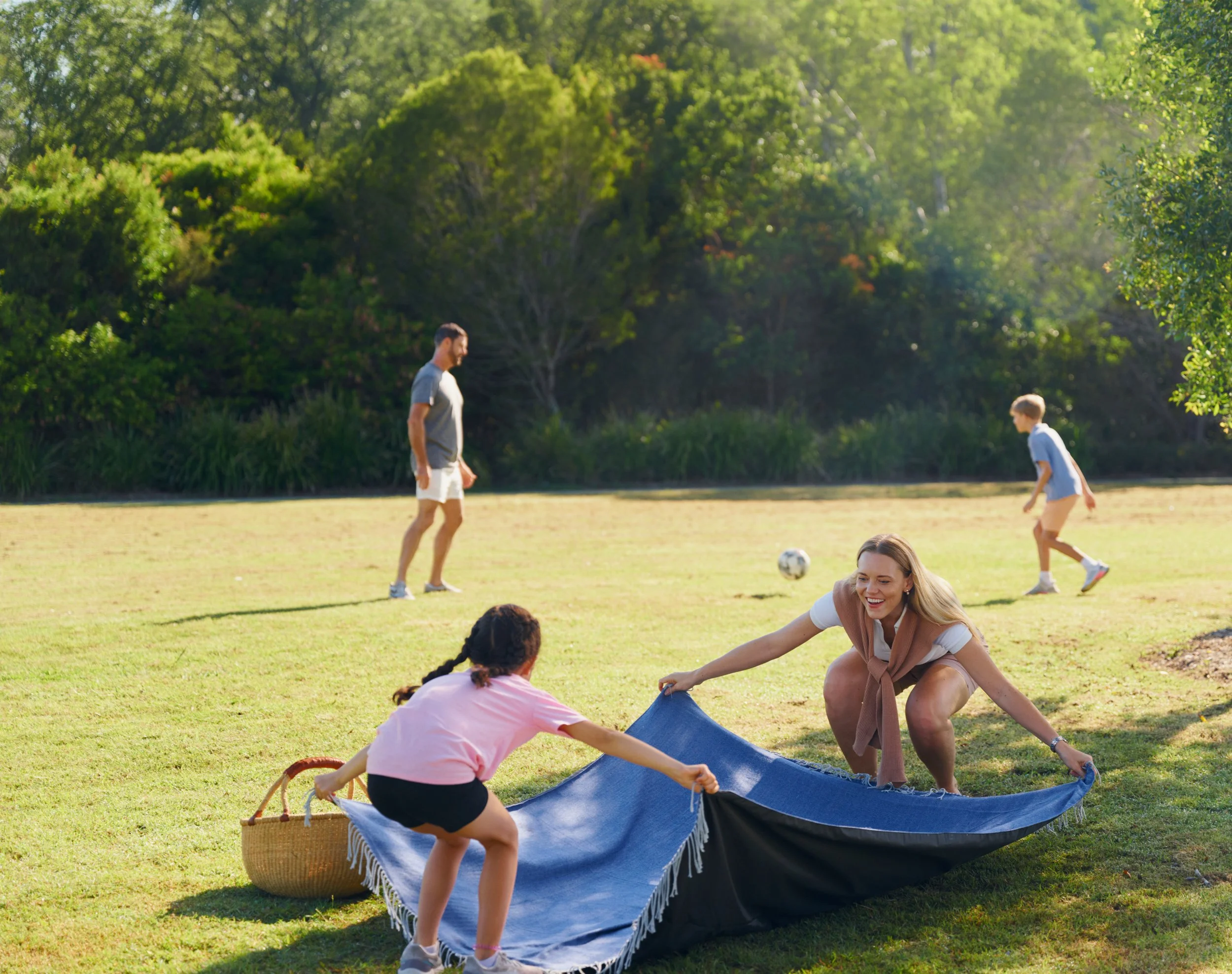 A diverse group of people playing tug-of-war with a blue cloth in a park with lush green trees in the background.