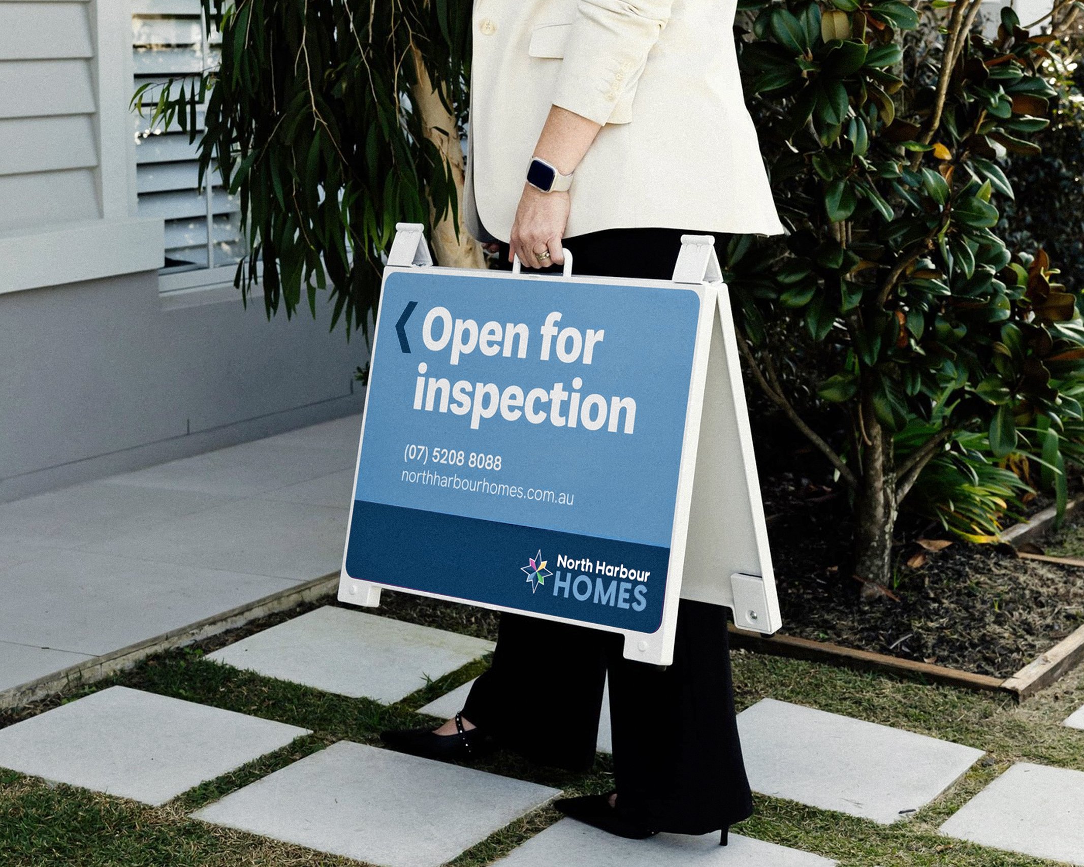 A person holding a sign that reads 'Open for inspection' outside a house surrounded by greenery.