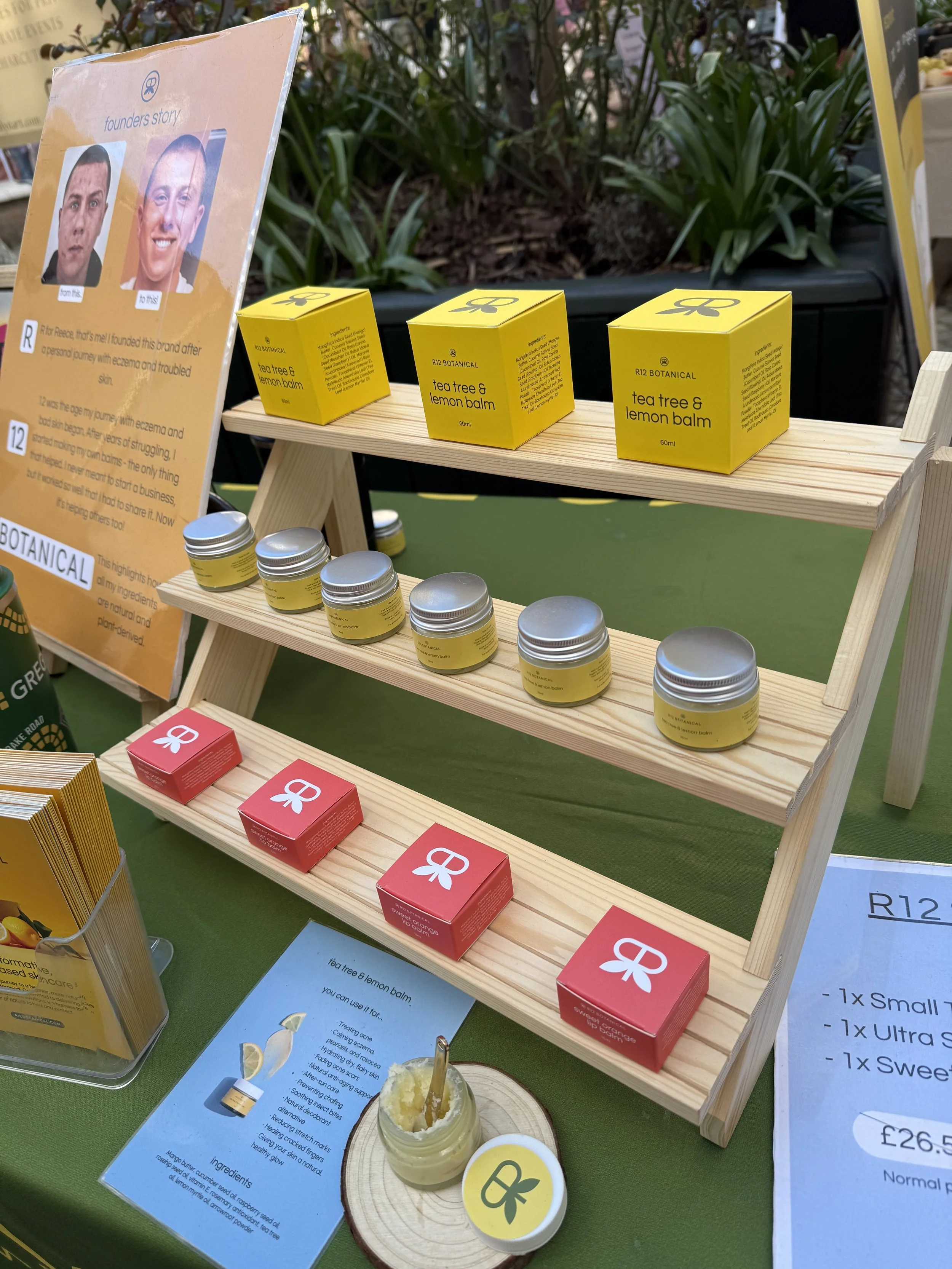 Display table featuring yellow boxes labeled 'tea tree & lemon balm' and small jars, with informational signs about ingredients and benefits of the products, and a poster illustrating a founder's story with before and after photos.