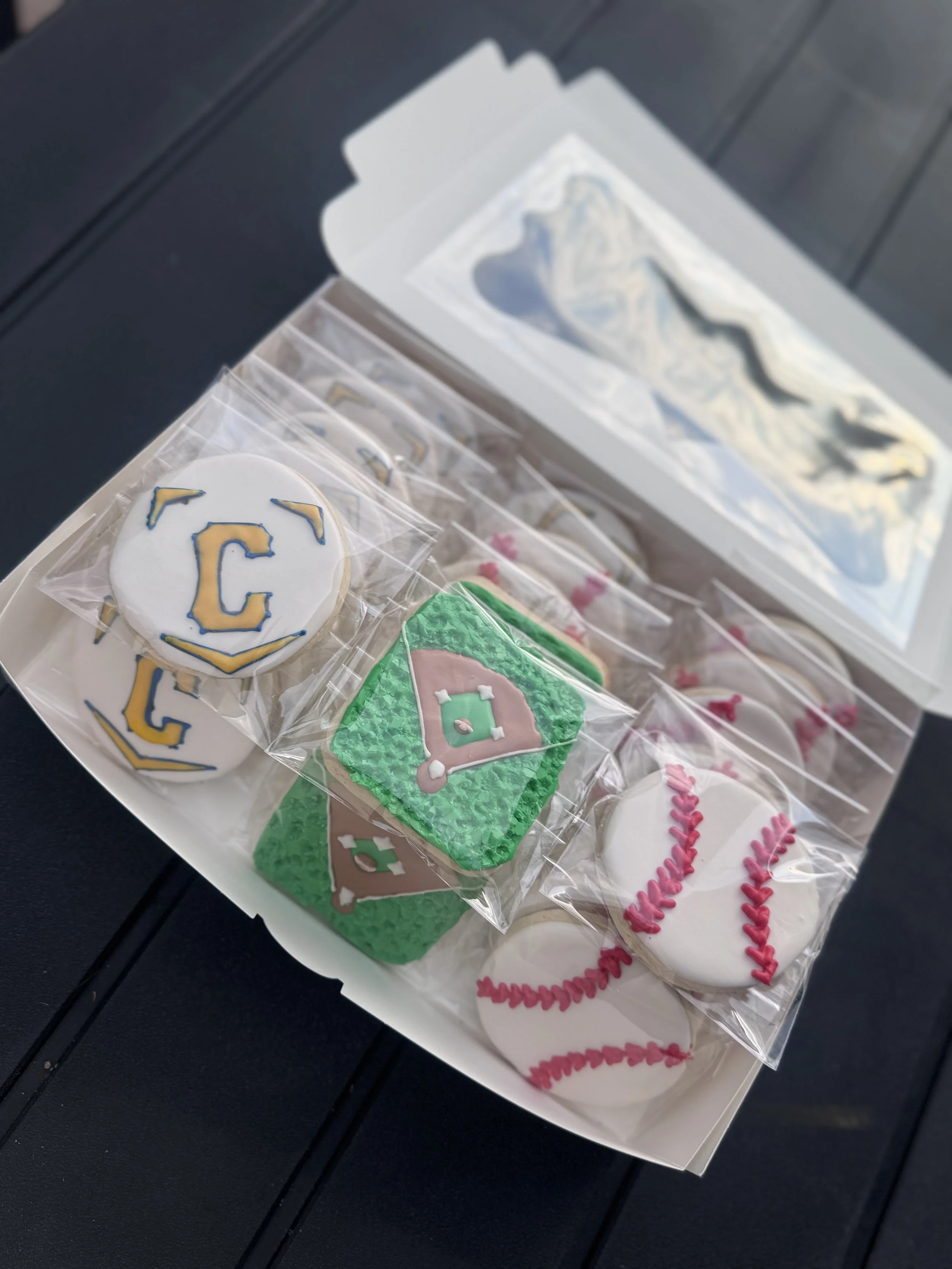 Box of baseball-themed cookies, decorated with baseballs, gloves, and team logos, wrapped in plastic and placed in a box with batting cage background.