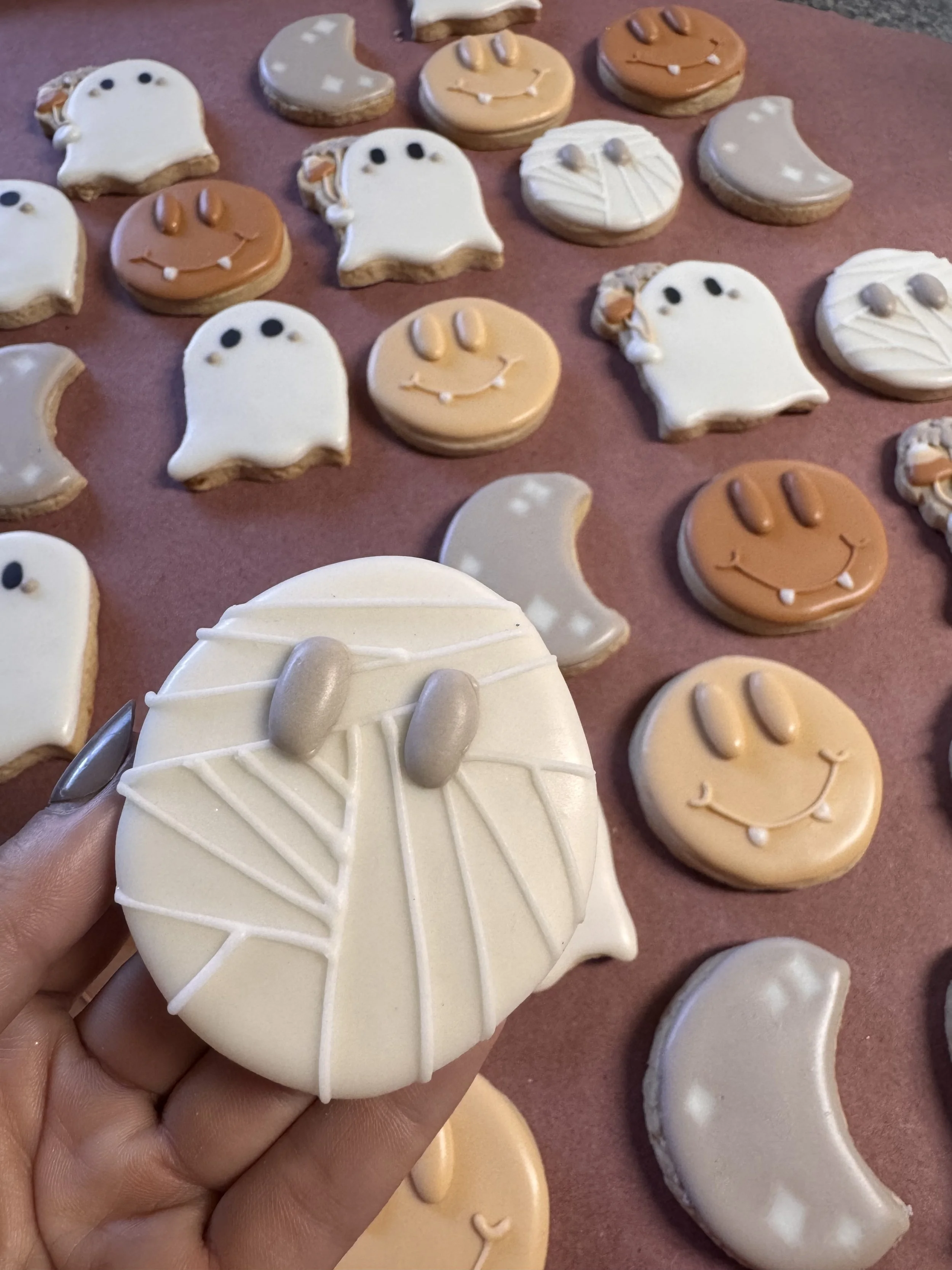 Assorted Halloween-themed cookies decorated with various icing designs, including ghosts, jack-o'-lanterns, crescent moons, and spiders on a pink surface.