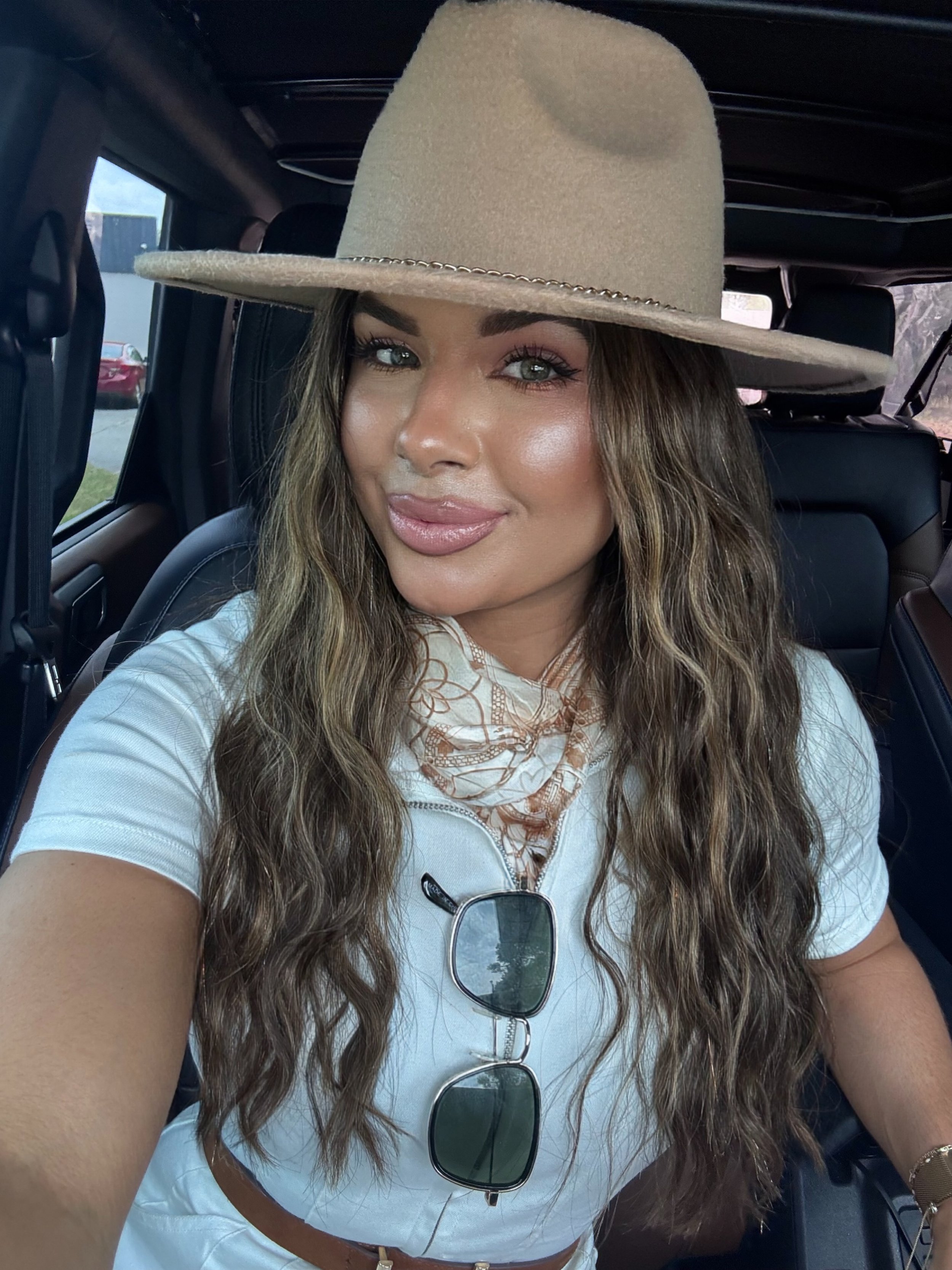 A woman with long wavy brown hair, wearing a beige wide-brimmed hat, white shirt, and sunglasses hanging from her shirt, sitting in a vehicle and taking a selfie.