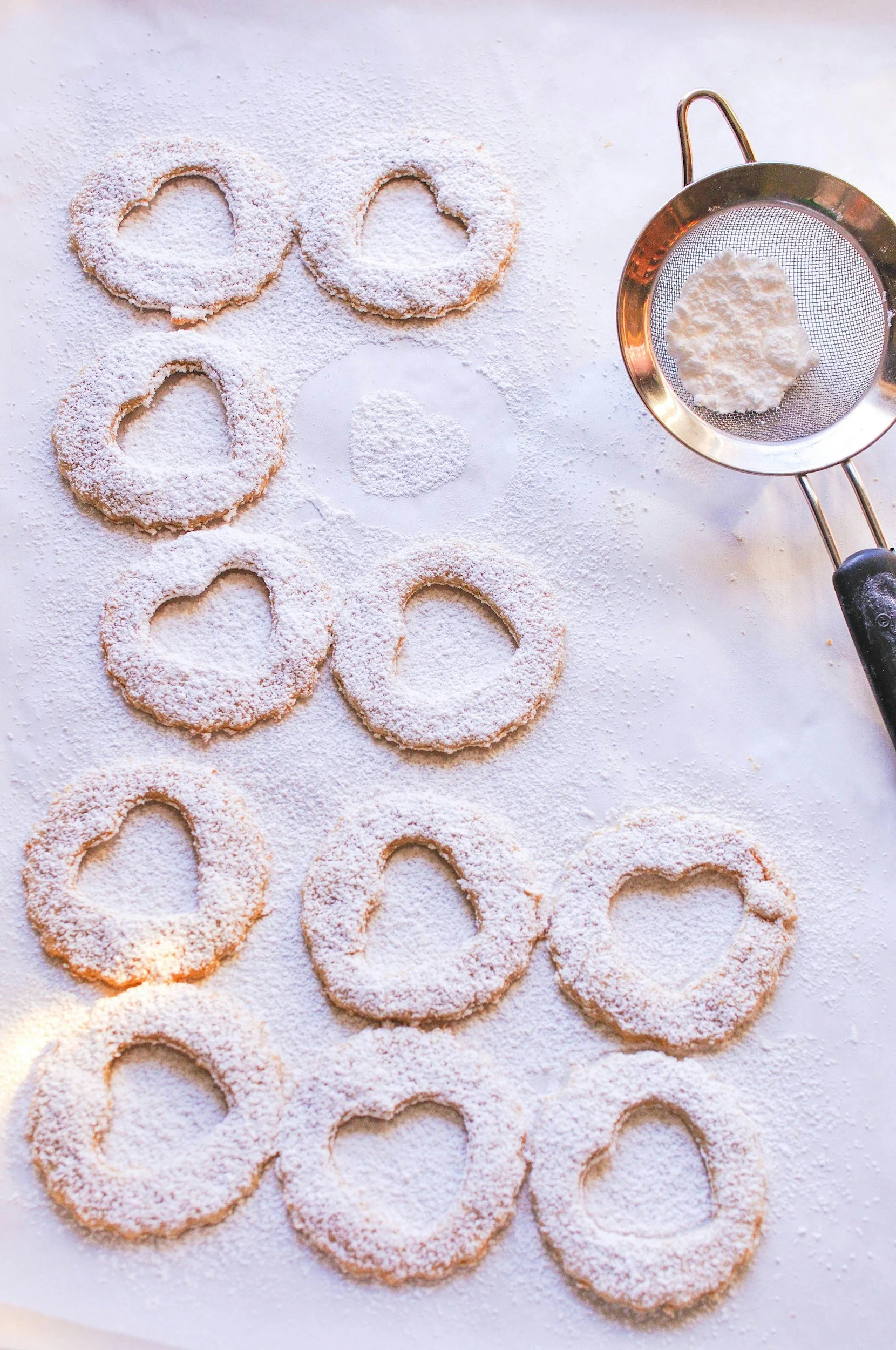 Heart-shaped cookies dusted with powdered sugar on a white surface, with a flour sifter and fork on the right.