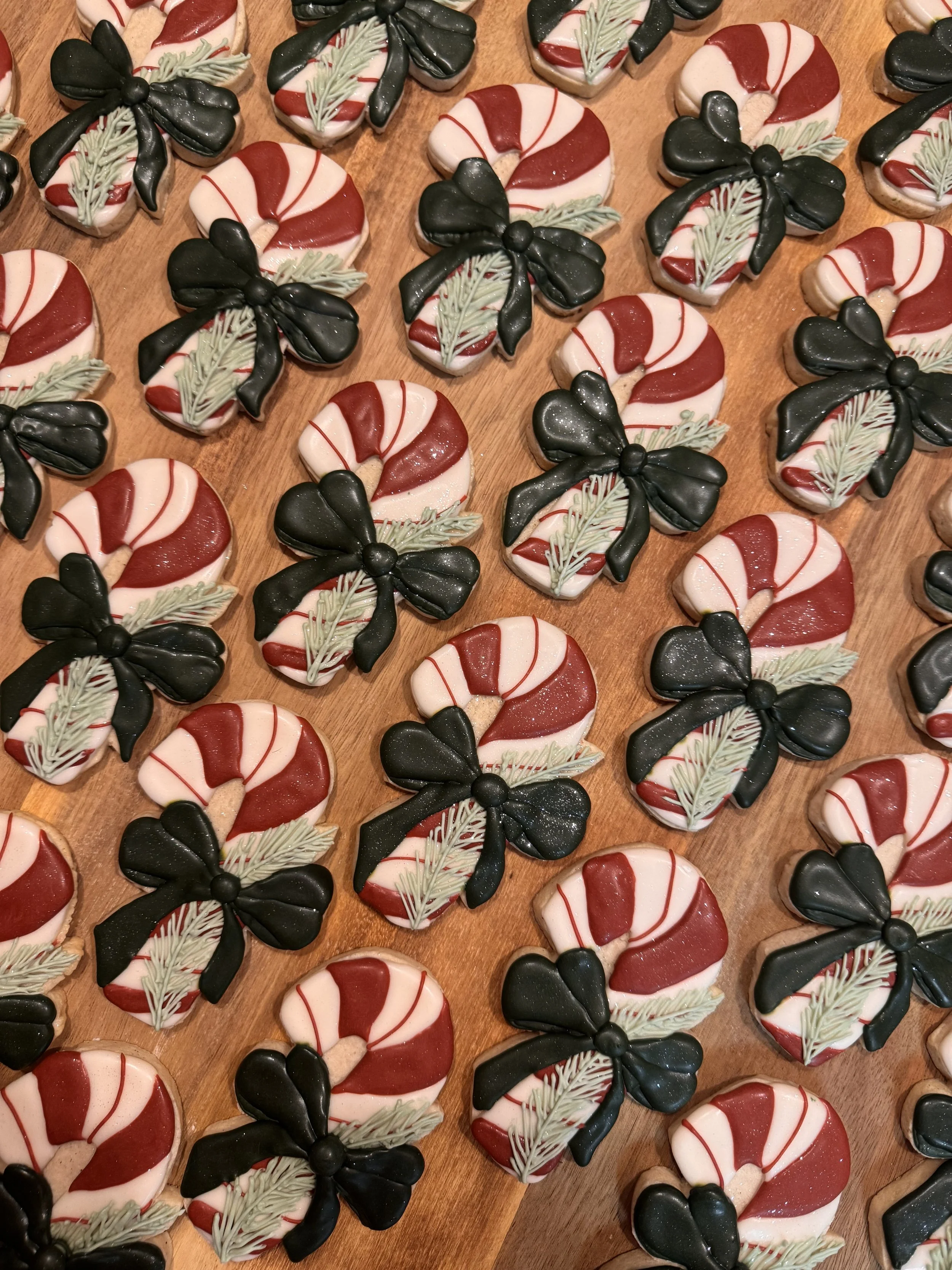 Holiday cookies shaped and decorated like Christmas candy canes with black bows and green festive accents, arranged on a wooden surface.