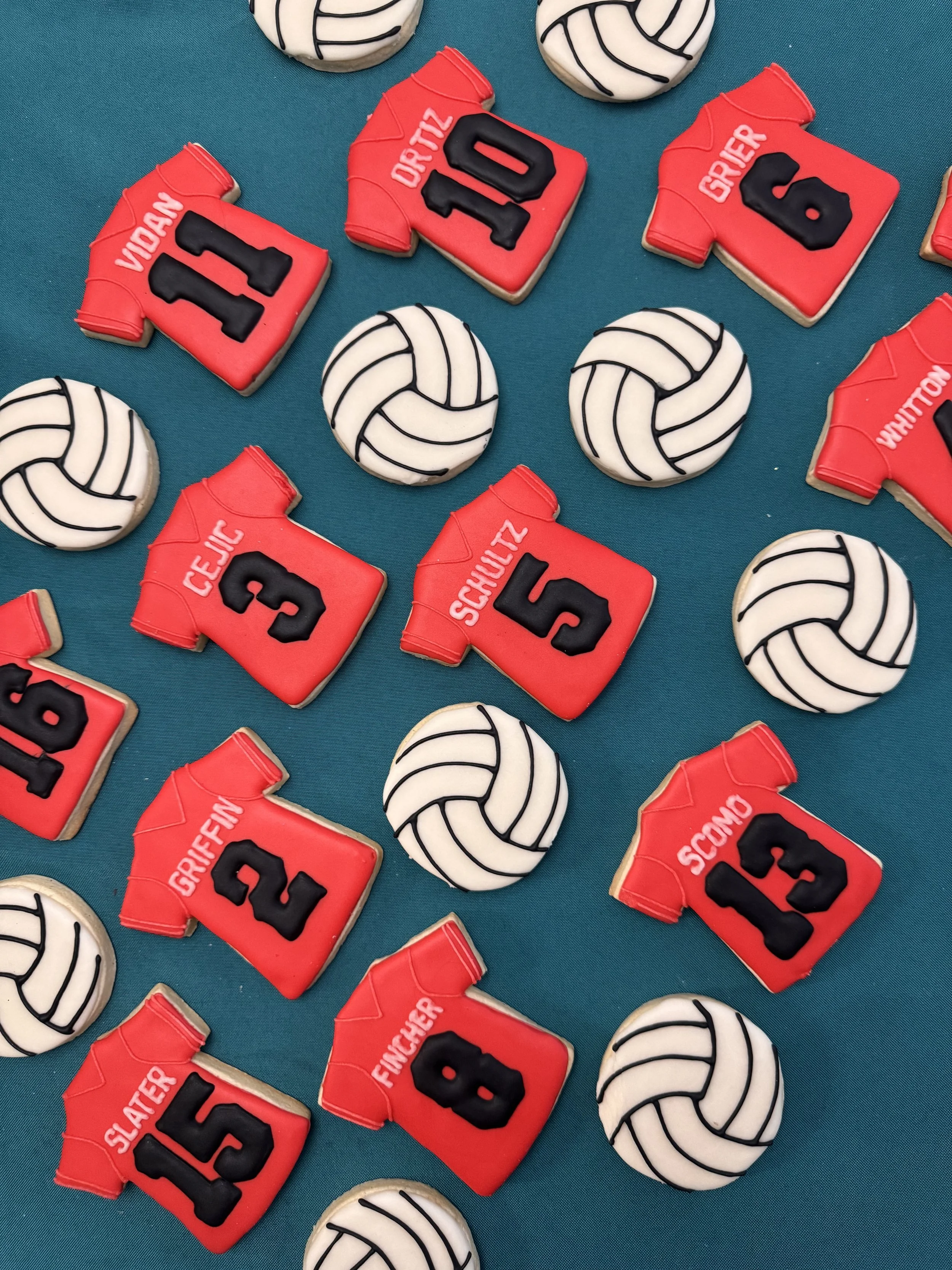 Decorative volleyball-themed cookies and jersey-shaped cookies with player names and numbers on a teal surface.