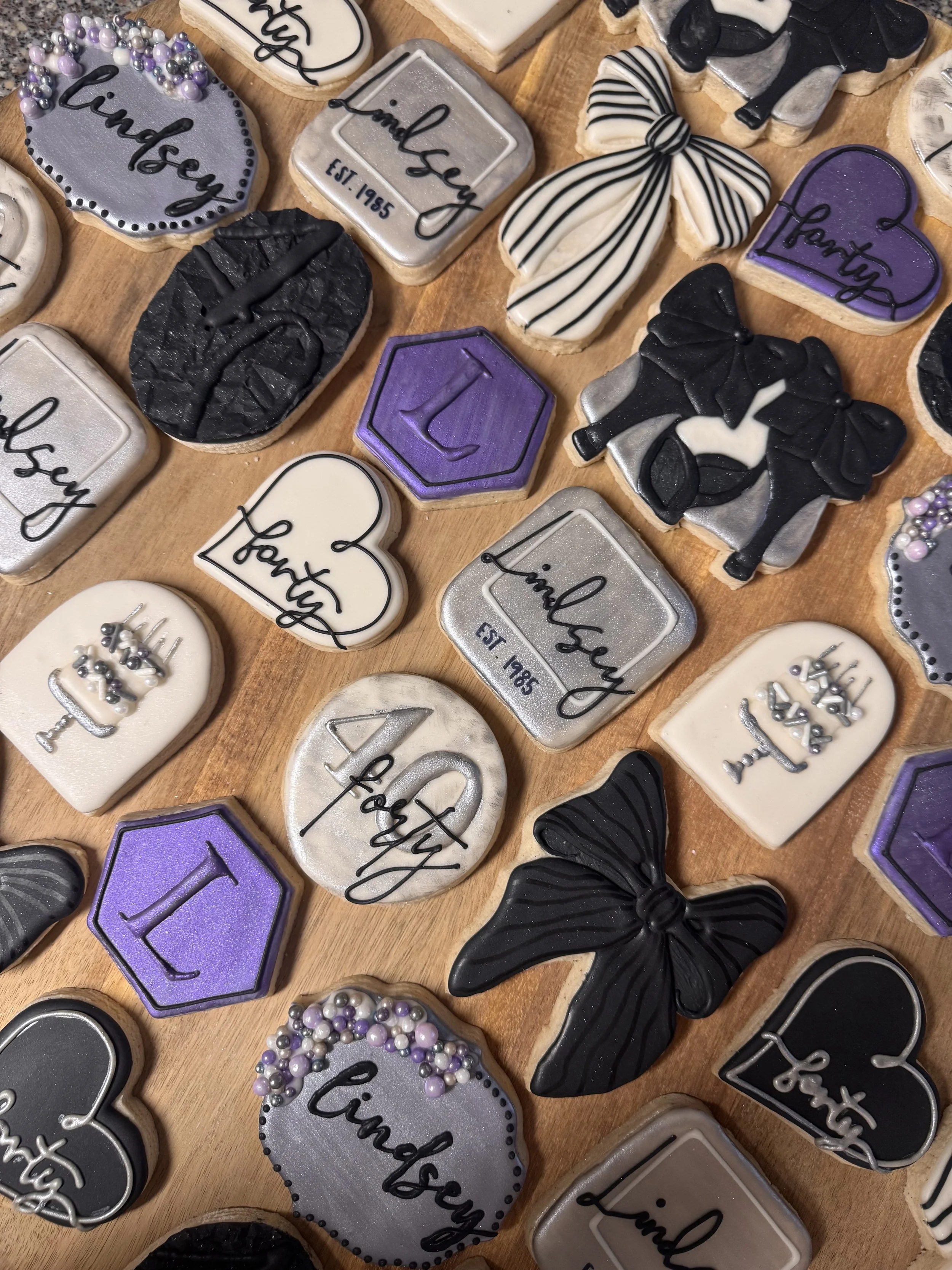 Decorated cookies with black, purple, silver, and white themes, featuring names, hearts, bows, and the number 40, arranged on a wooden surface.