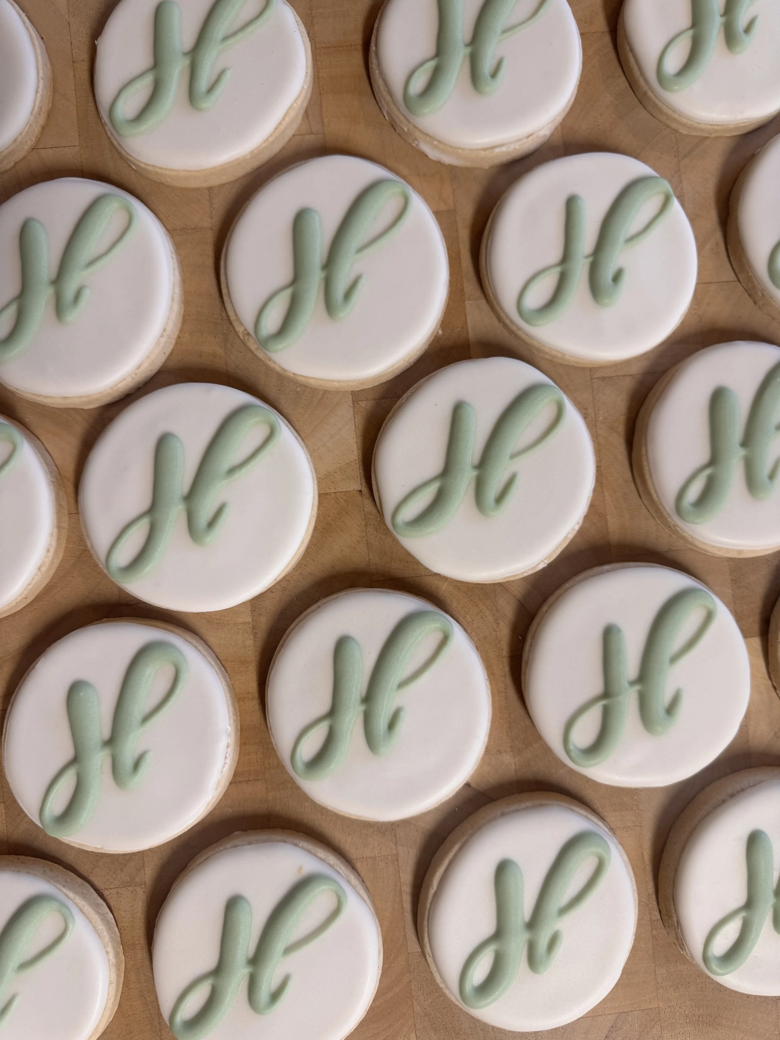 Round cookies with white icing and a light green letter 'H' piped on top, arranged on a wooden surface.