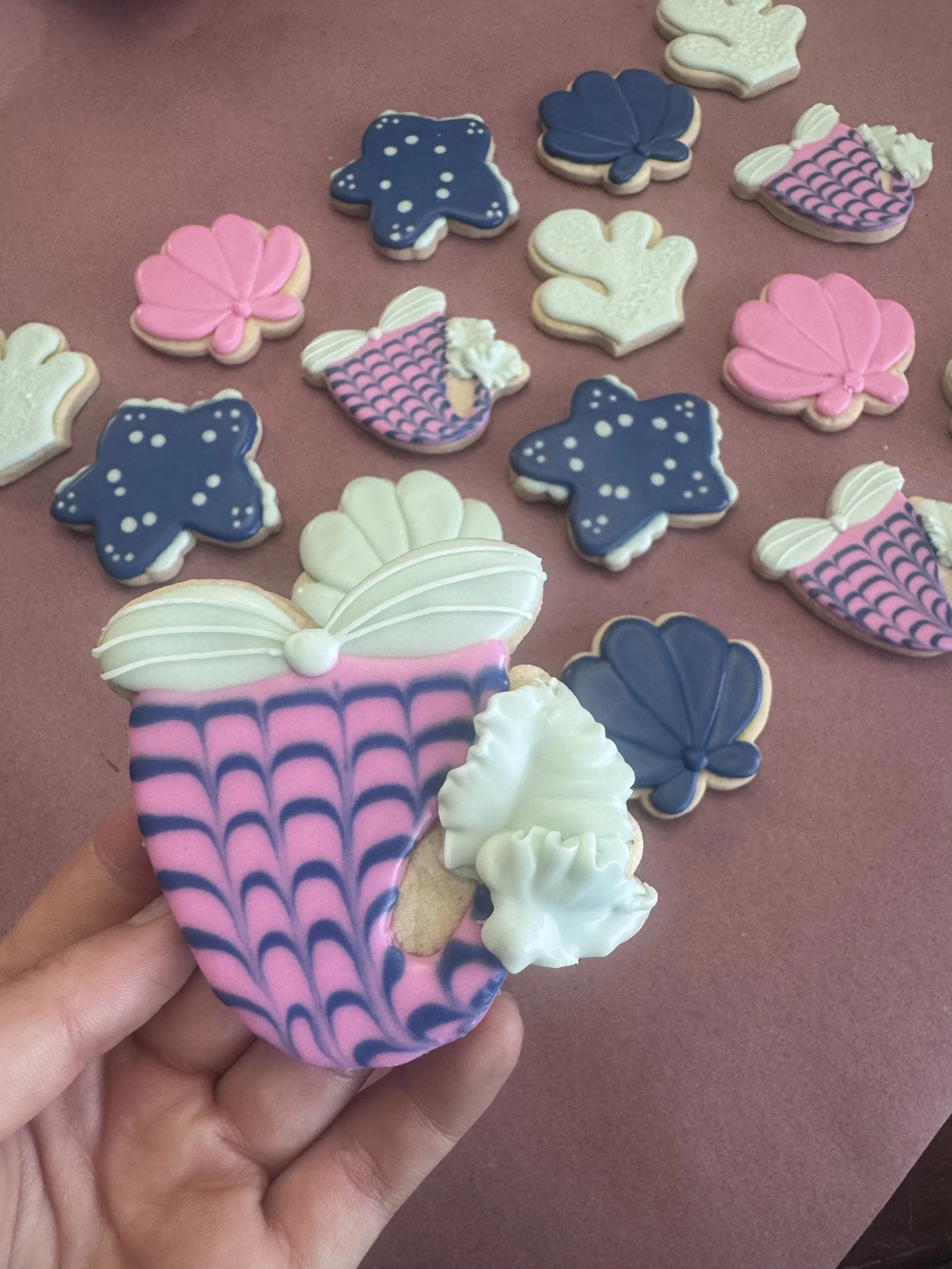 Decorative cookies in butterfly, flower, and seashell shapes with pink, blue, white, and purple icing, laid out on a pink surface, with one cookie held in hand featuring a pink and blue pattern.